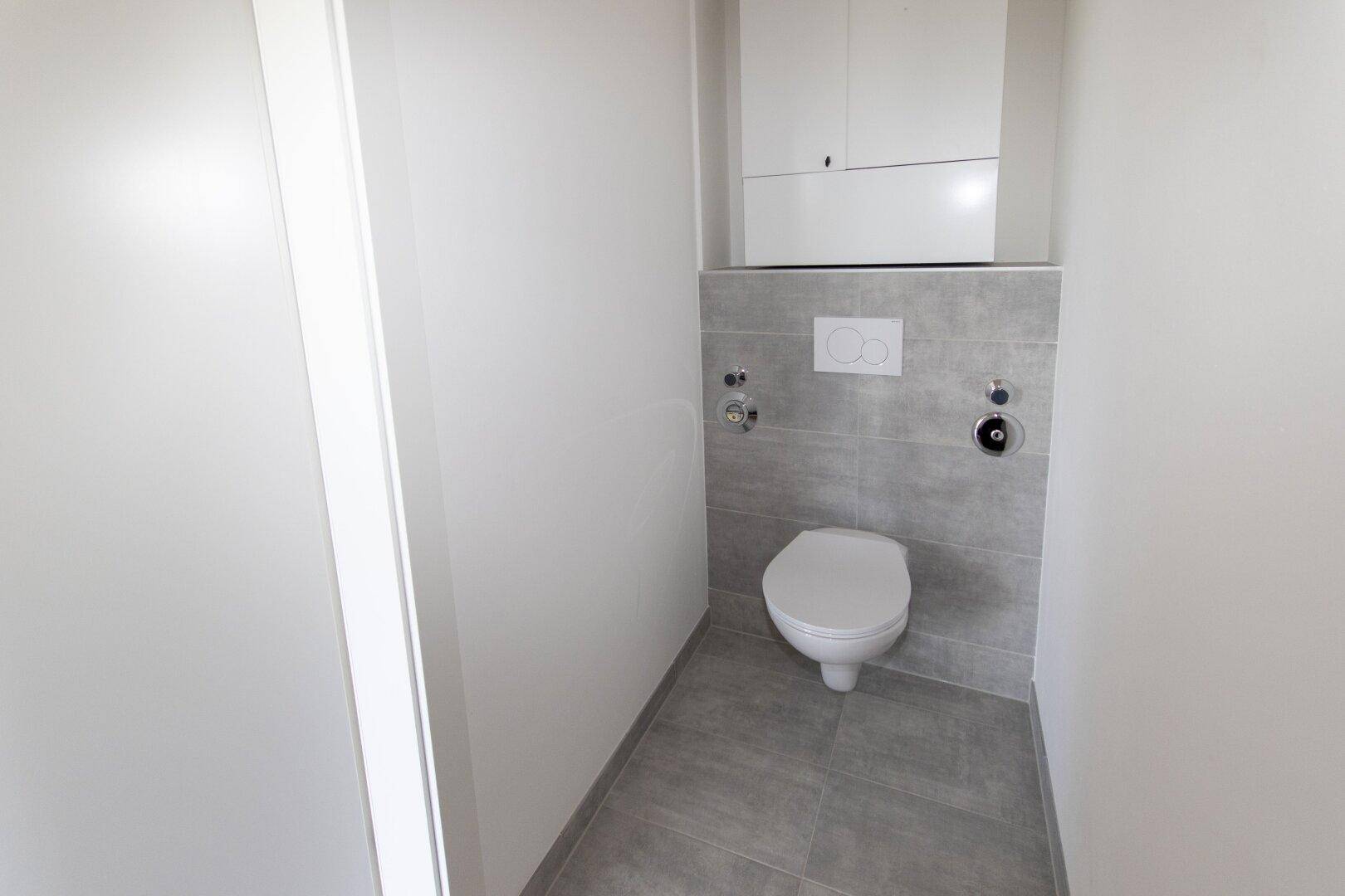 Separates WC mit wandmontierter Toilette und modernen grauen Fliesen an Wand und Boden.