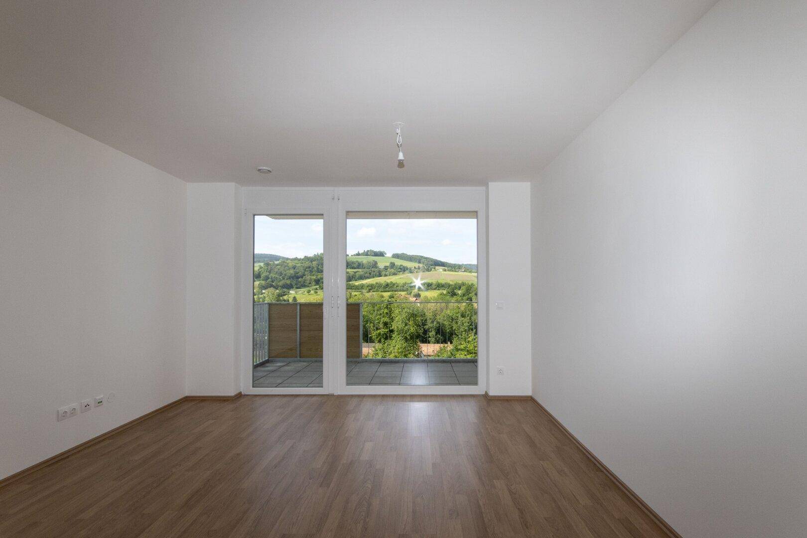 Lichtdurchfluteter Wohnbereich mit Holzboden und weitem Blick ins Grüne durch die Balkontür.