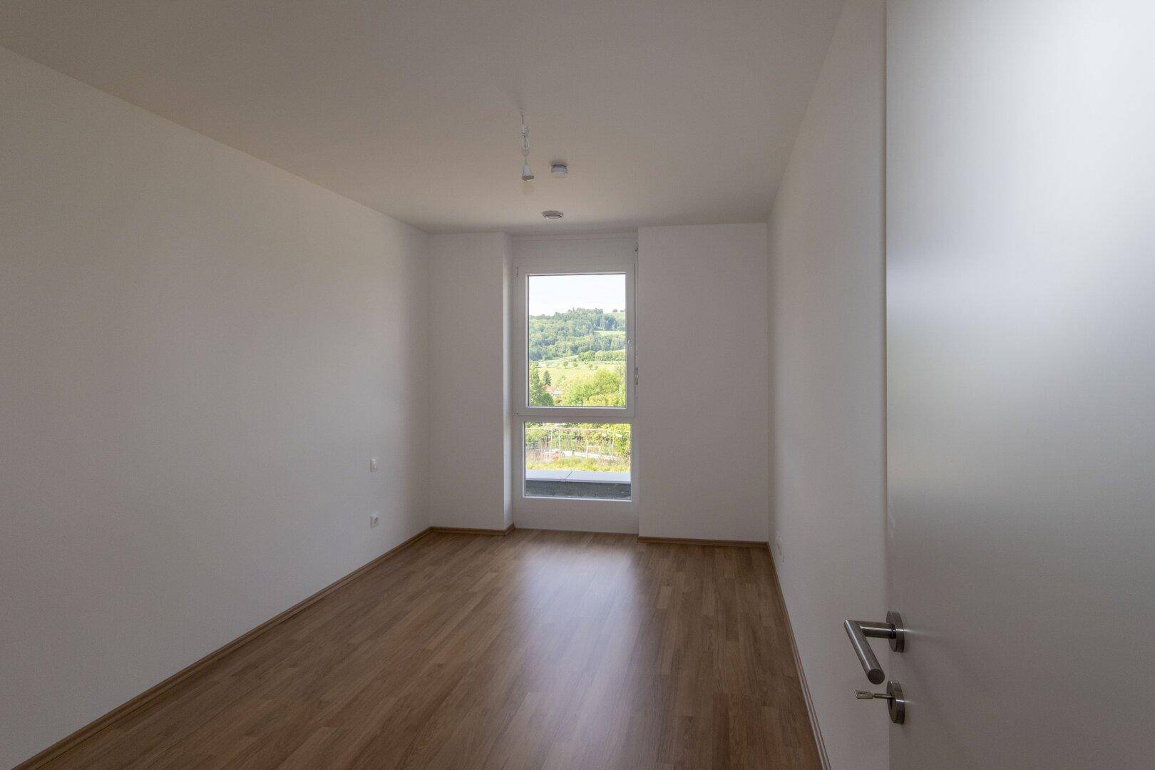 Geräumiges Schlafzimmer mit Holzboden und Fenster, das viel Tageslicht und eine grüne Aussicht bietet.