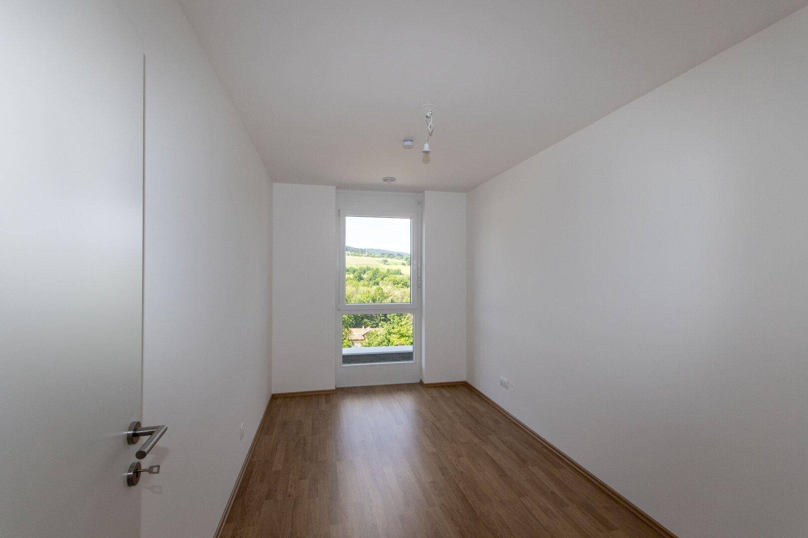 Helles Schlafzimmer mit Holzboden und großem Fenster, das einen schönen Blick ins Grüne bietet.