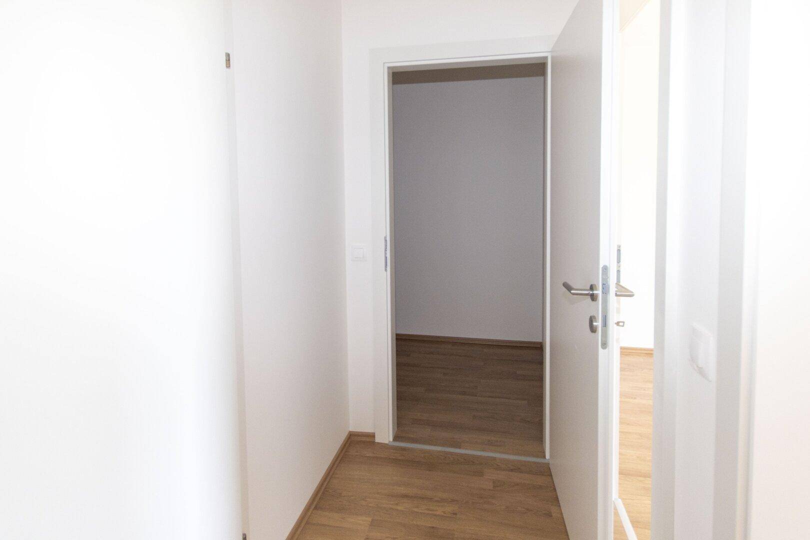 Heller Flur mit Holzboden und offener Tür, die den Blick in ein weiteres Zimmer freigibt.