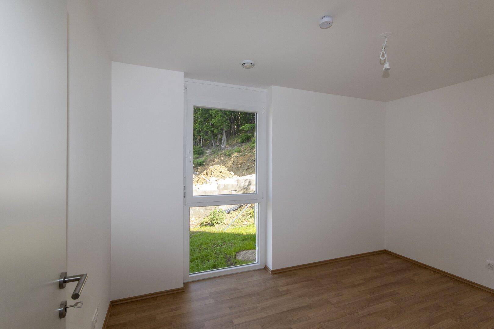 Helles Schlafzimmer mit Laminatboden und Fenster mit Blick auf eine grüne Böschung.