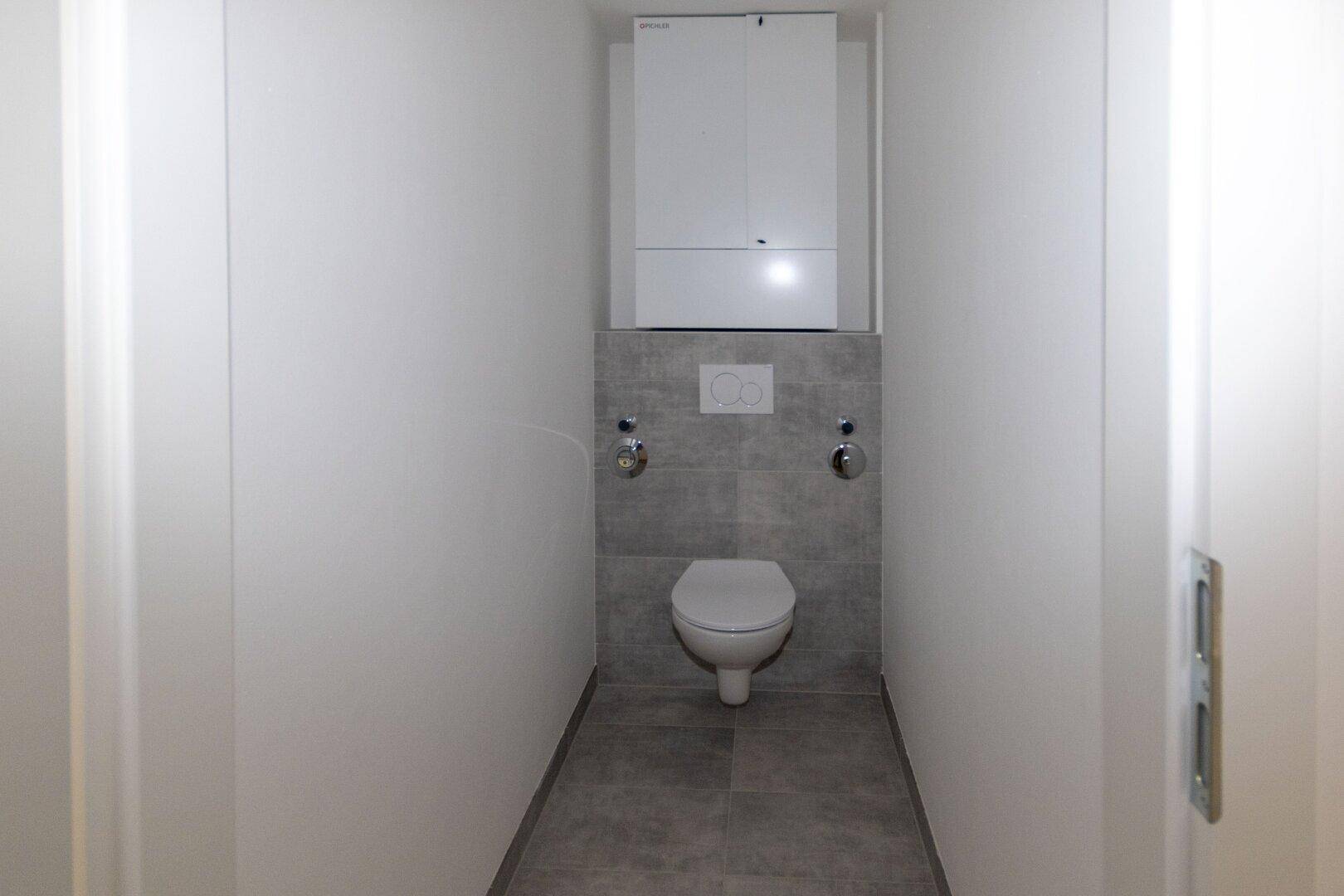 Separates WC mit wandhängender Toilette, grauen Fliesen und praktischen Oberschränken.
