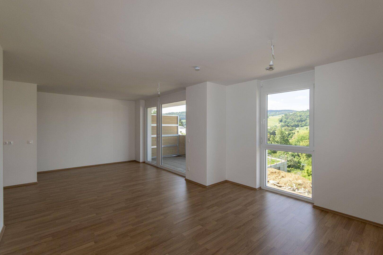 Helles Wohnzimmer mit Laminatboden, weißen Wänden und großen Fenstern mit Blick ins Grüne.