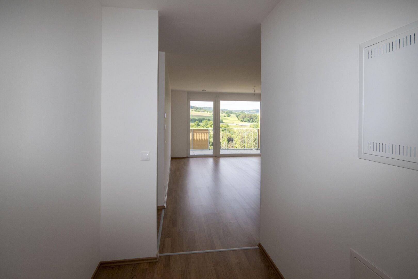 Langer, heller Flur mit Laminatboden, der zum Wohnbereich mit Balkon und Aussicht führt.