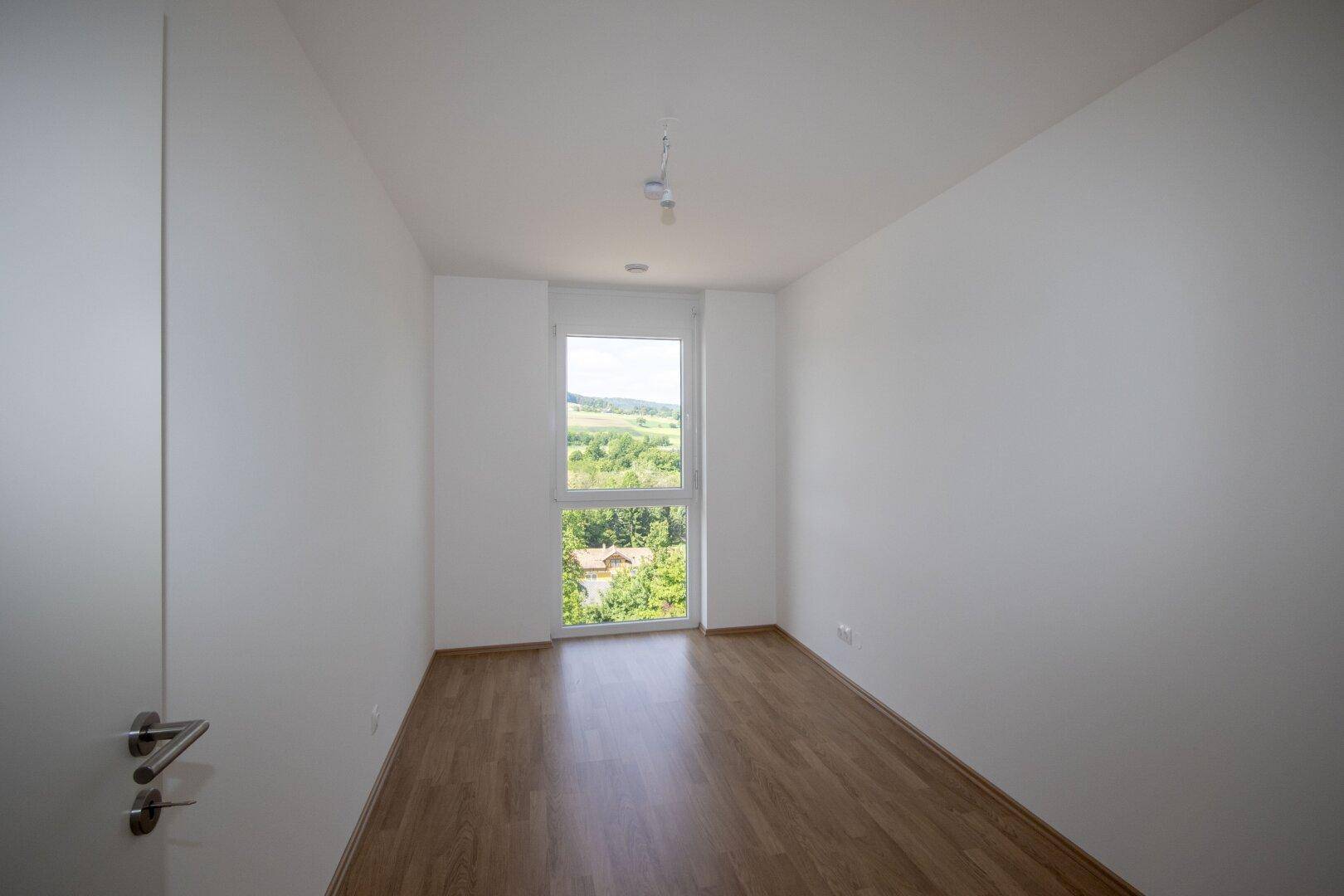 Helles Schlafzimmer mit Laminatboden und einem Fenster, das einen schönen Blick ins Grüne bietet.