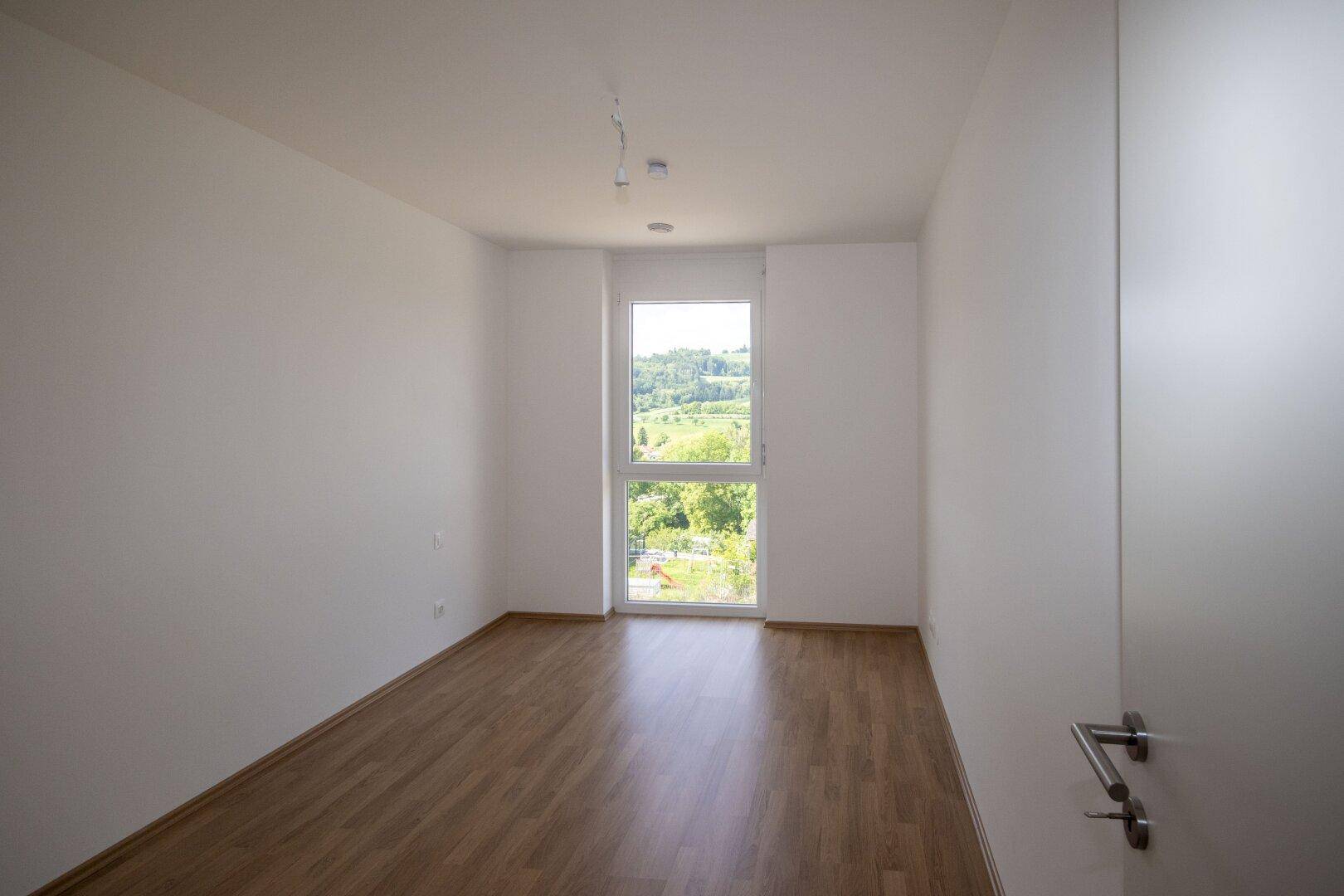 Freundliches Zimmer mit Laminatboden und großem Fenster, das viel Tageslicht und eine grüne Aussicht bietet.