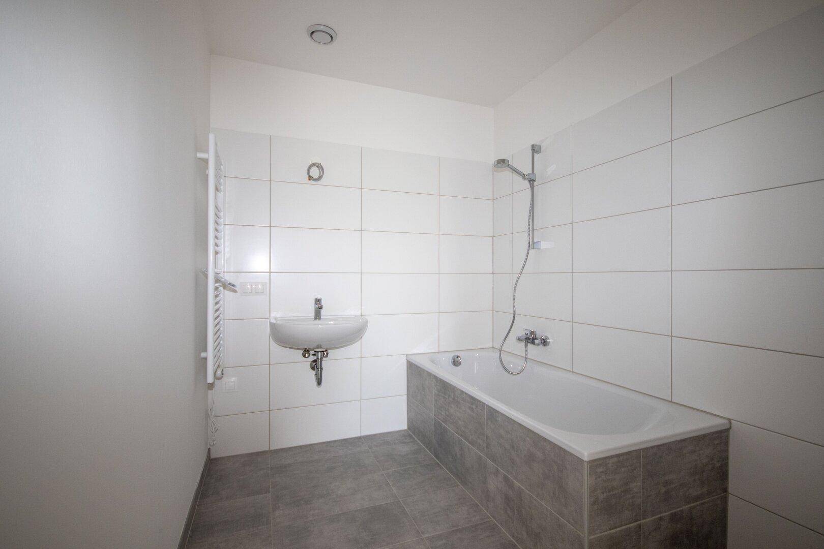 Modernes Badezimmer mit weißen Wandfliesen, grauen Bodenfliesen, Badewanne und Handtuchheizkörper.