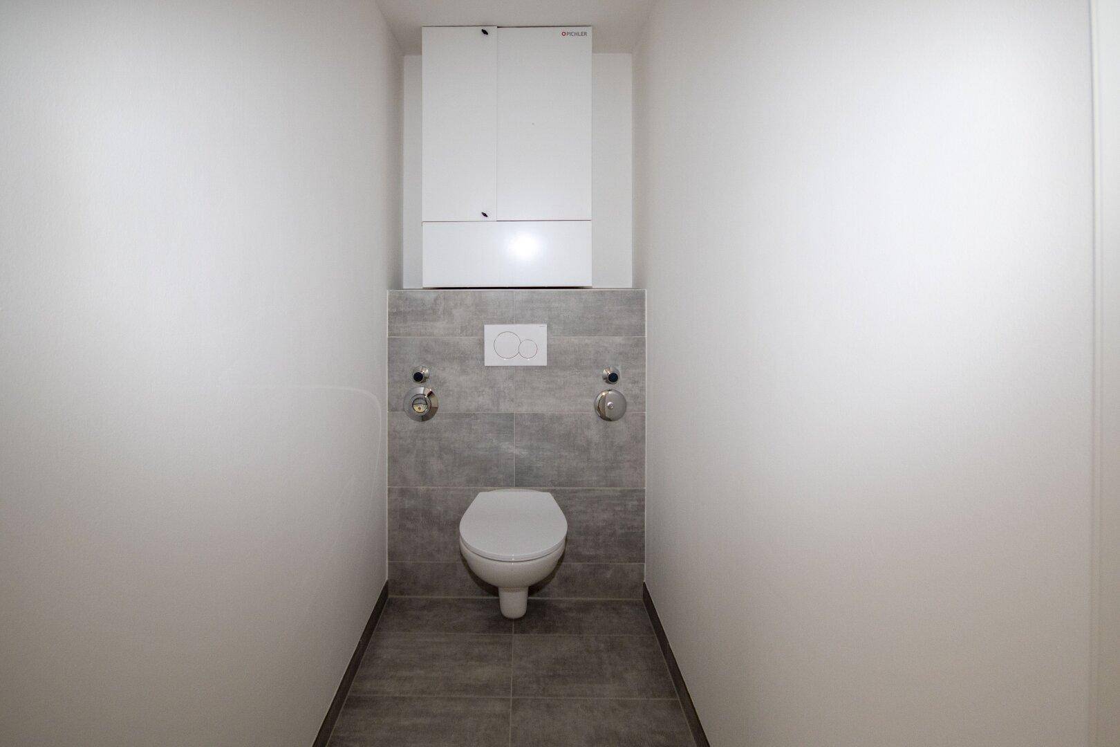 Separates WC mit modernen grauen Fliesen, wandhängender Toilette und praktischem Wandschrank.