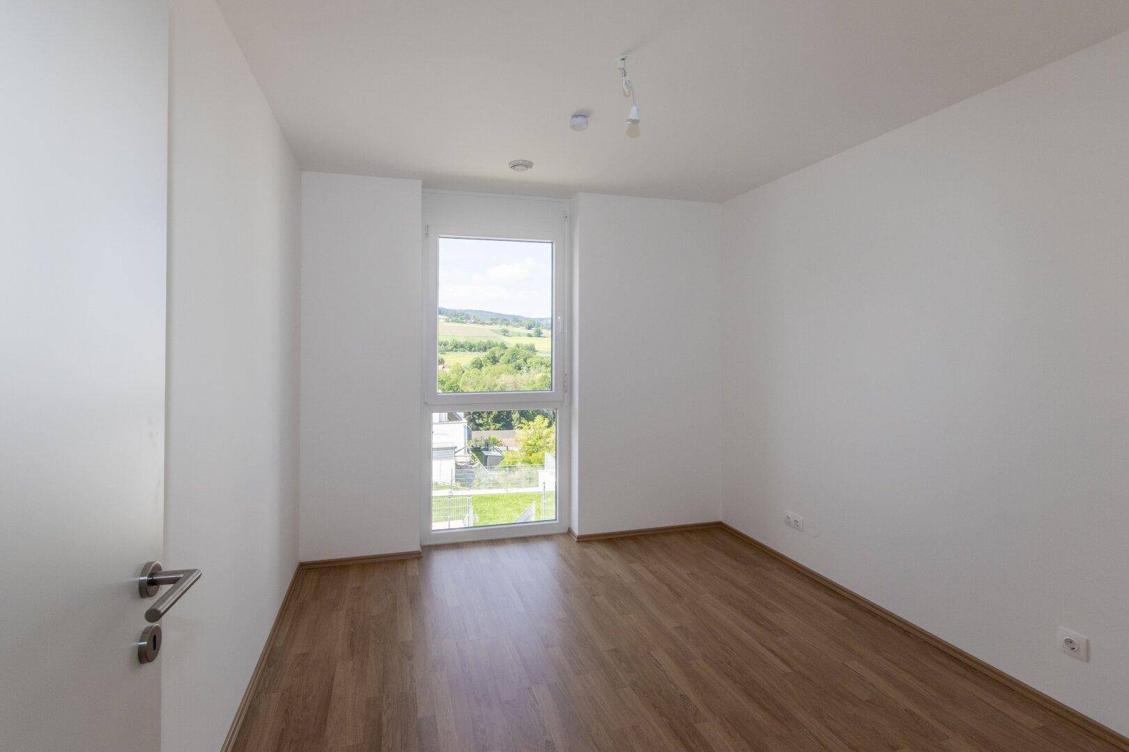 Helles Schlafzimmer mit Holzboden und großem Fenster, das viel Tageslicht hereinlässt.