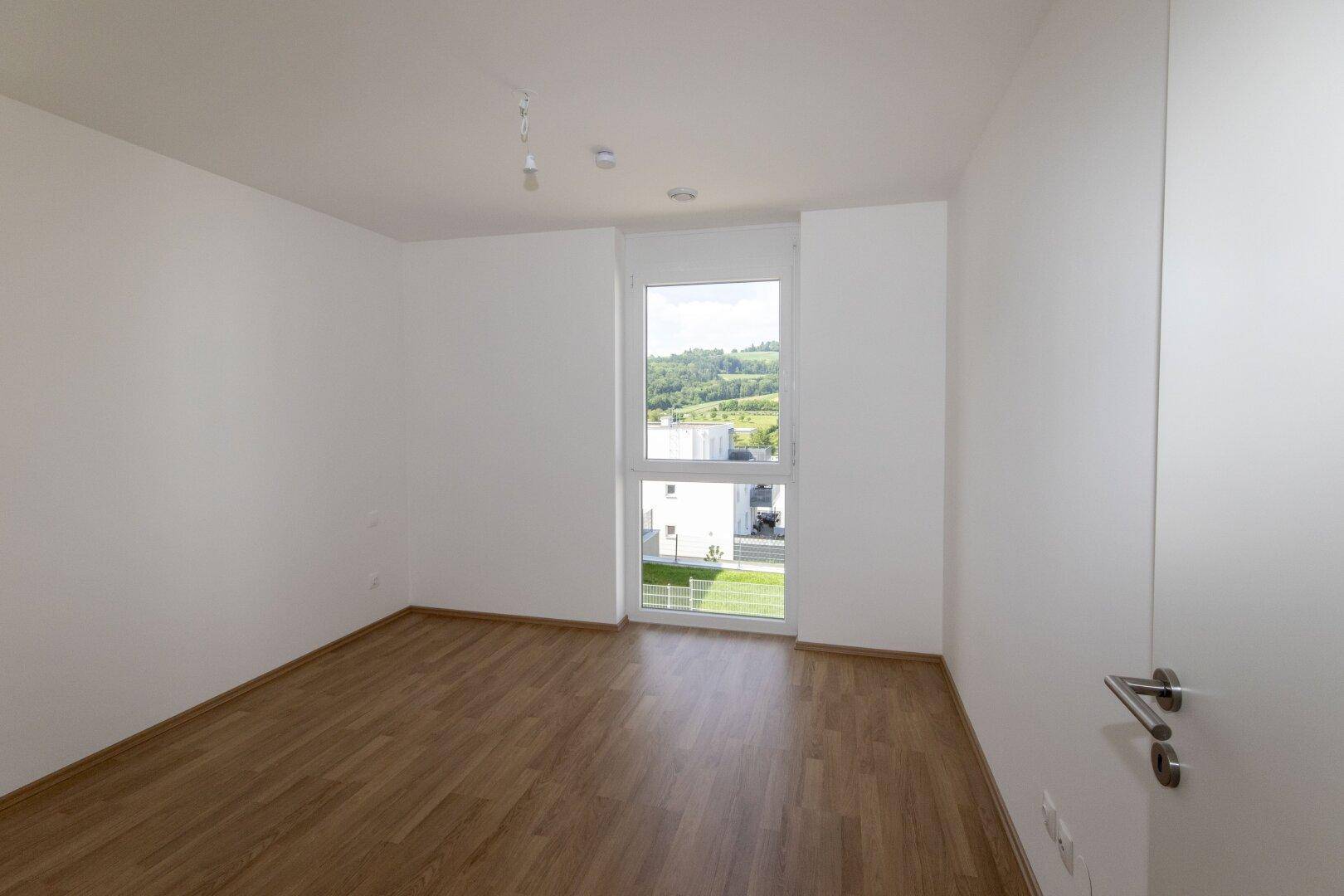 Leeres Schlafzimmer mit Holzboden und großem Fenster mit Blick auf die Umgebung.