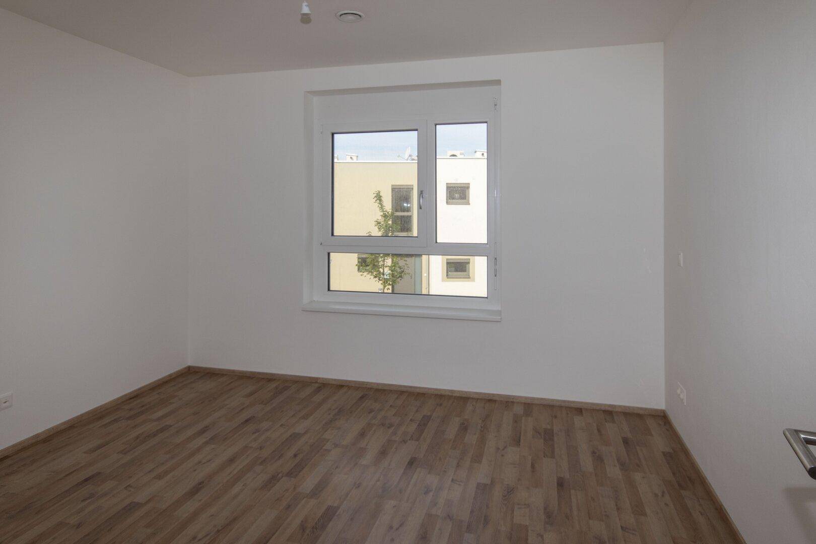 Helles Schlafzimmer mit Holzboden und Fenster mit Blick auf die umliegenden Gebäude.