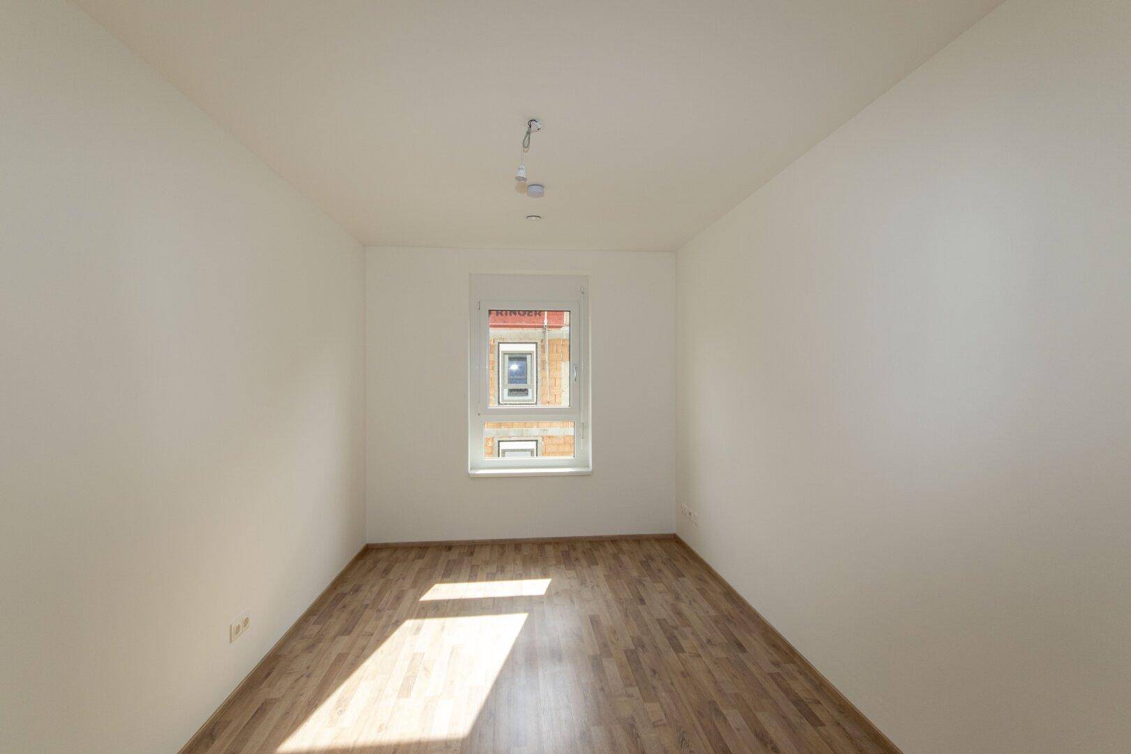 Helles Schlafzimmer mit Laminatboden und einem Fenster mit Blick auf ein Nachbargebäude.