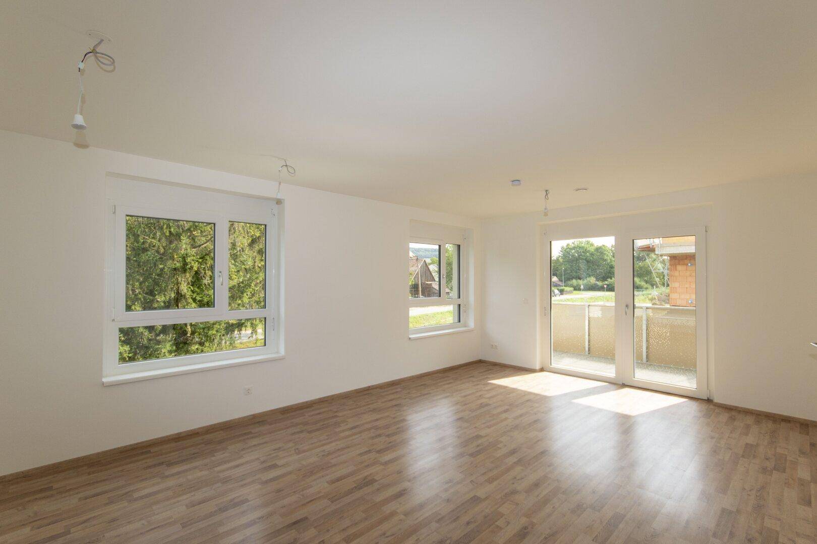 Helles, leeres Zimmer mit Laminatboden, weißen Wänden und Zugang zu einem Balkon.