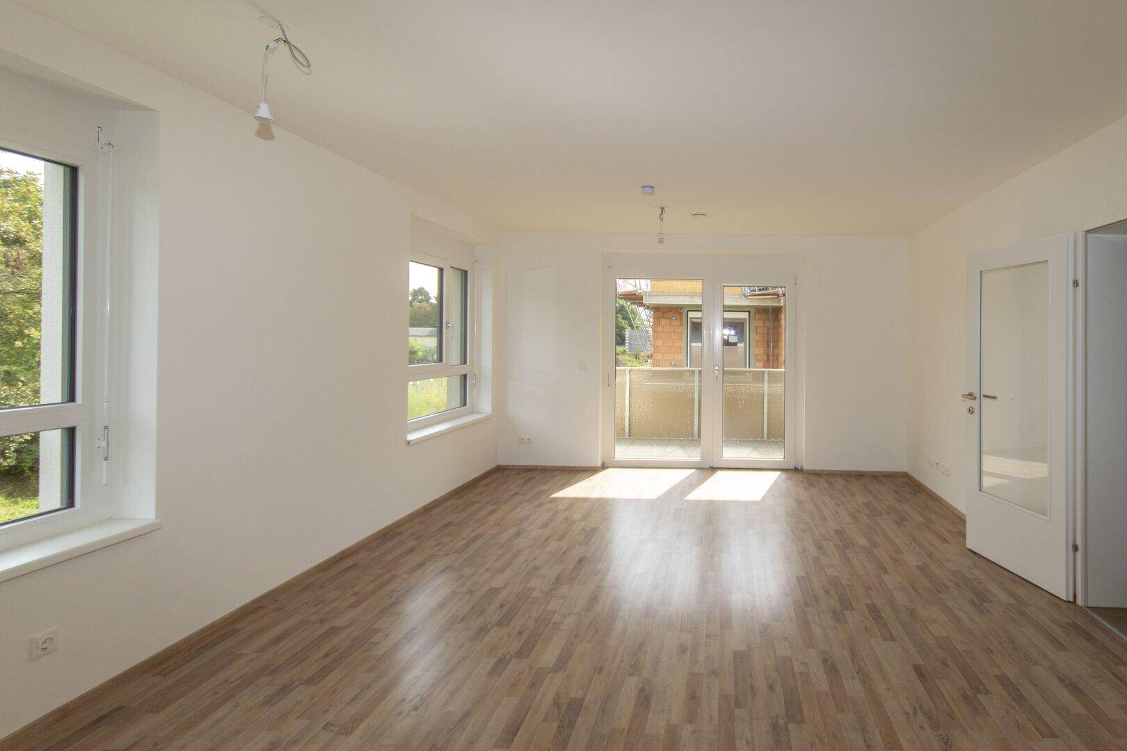 Geräumiges, helles Zimmer mit Laminatboden, weißen Wänden und Balkonzugang.