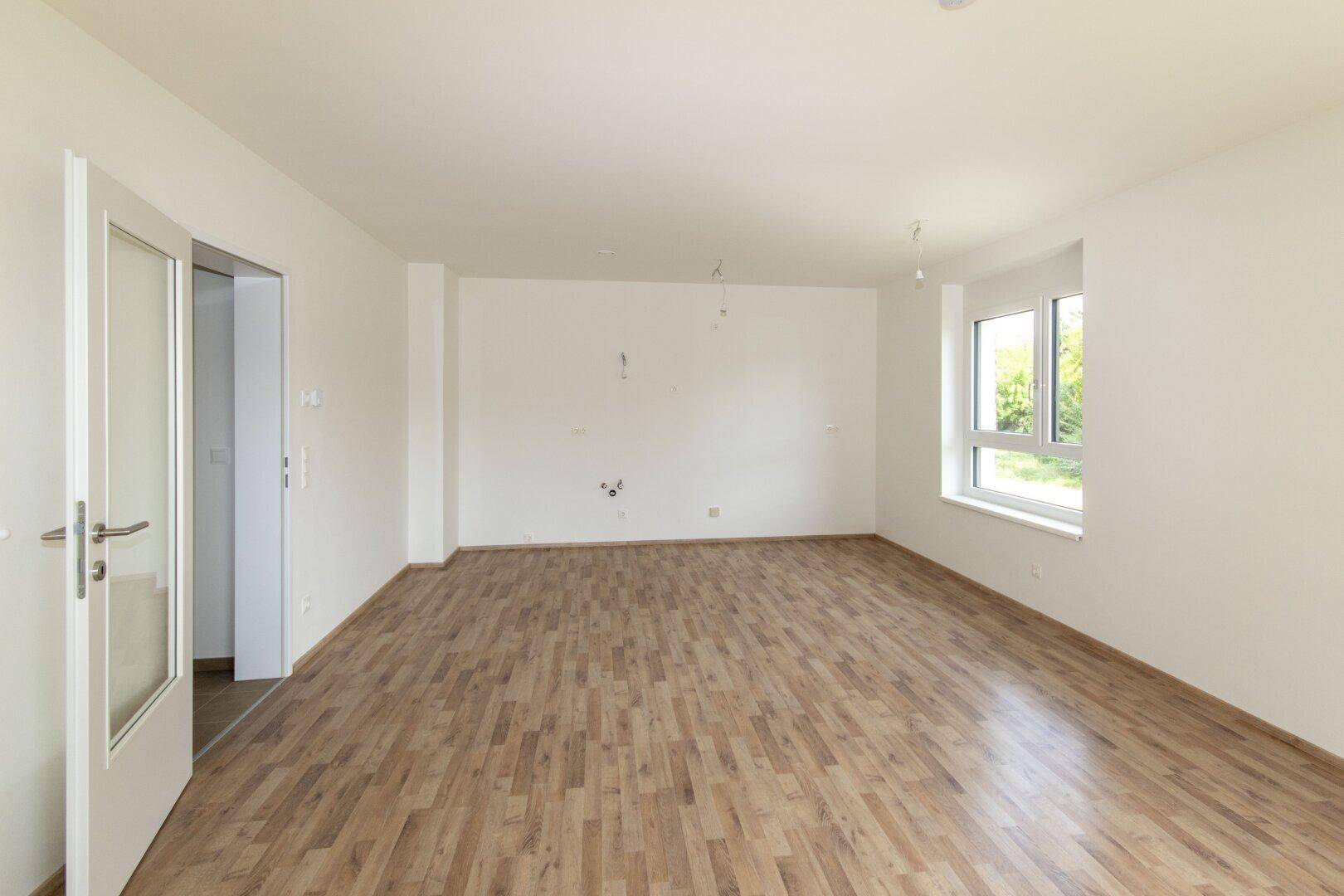 Leeres Zimmer mit hellem Laminatboden, weißen Wänden und einem Fenster mit Blick nach draußen.