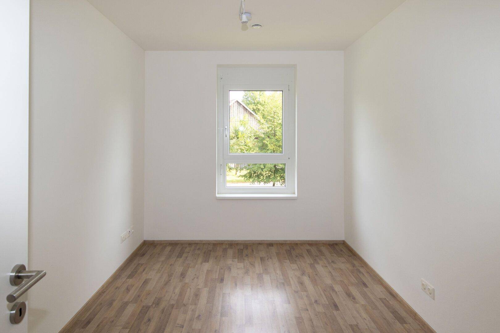 Ruhiges Schlafzimmer mit hellem Laminatboden und einem Fenster, das viel Tageslicht hereinlässt.
