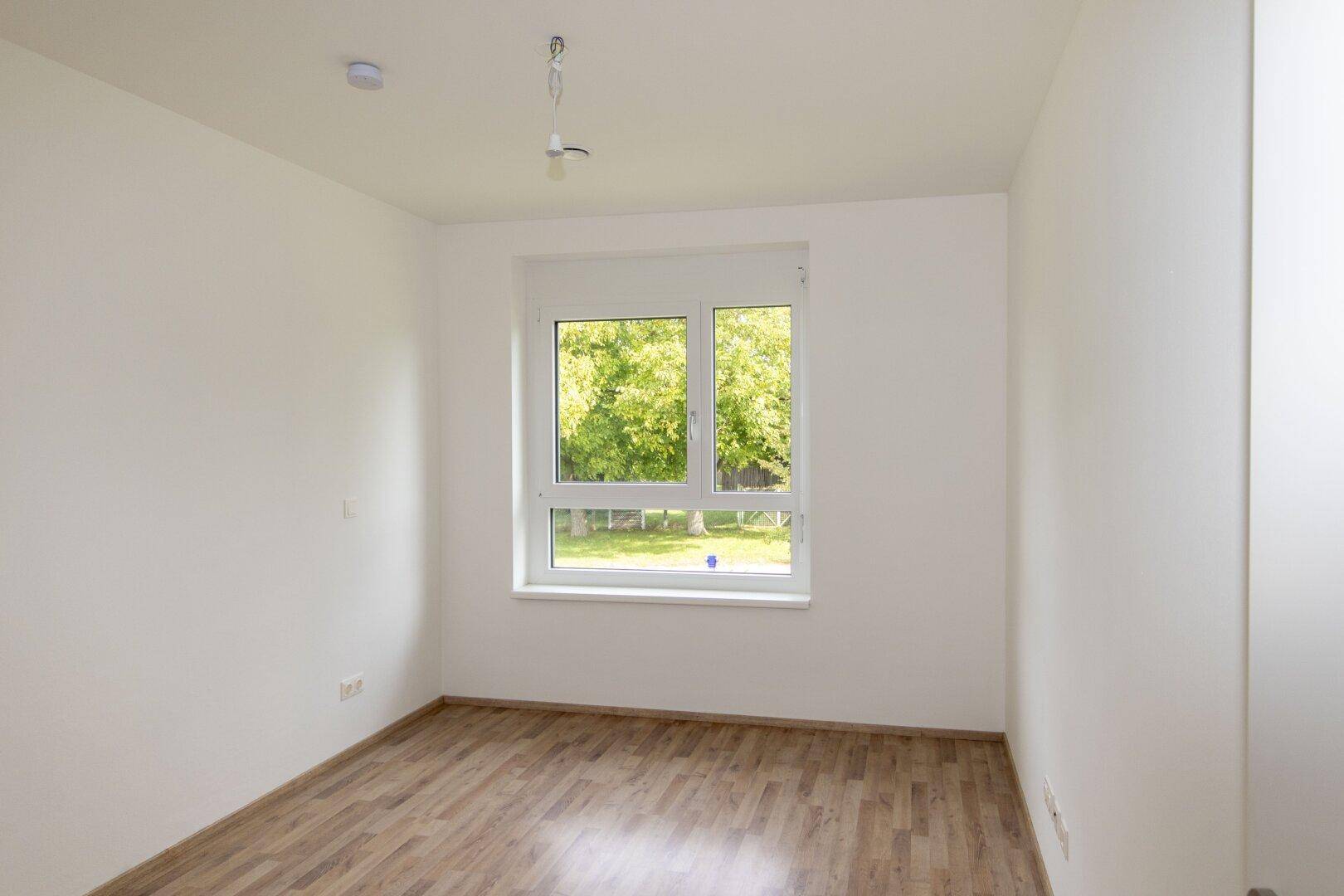 Helles Schlafzimmer mit Laminatboden und einem großen Fenster, das einen angenehmen Blick ins Grüne bietet.