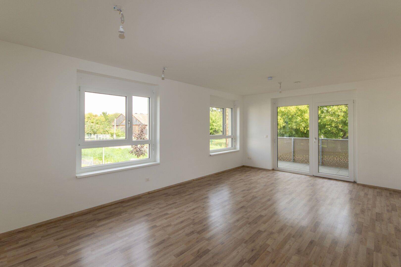Helles, leeres Wohnzimmer mit modernem Laminatboden und großen Fenstern mit Balkonzugang.