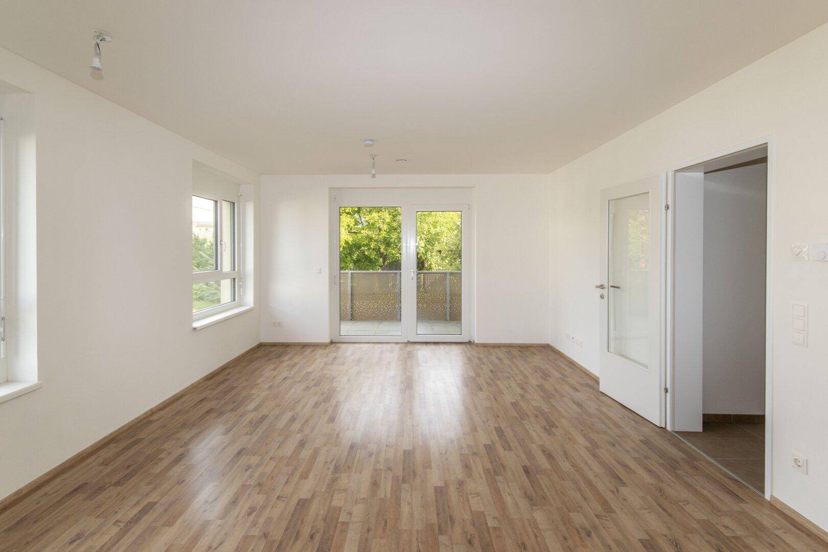Geräumiges Wohnzimmer mit hellem Laminatboden, weißen Wänden und Zugang zu weiteren Räumen.