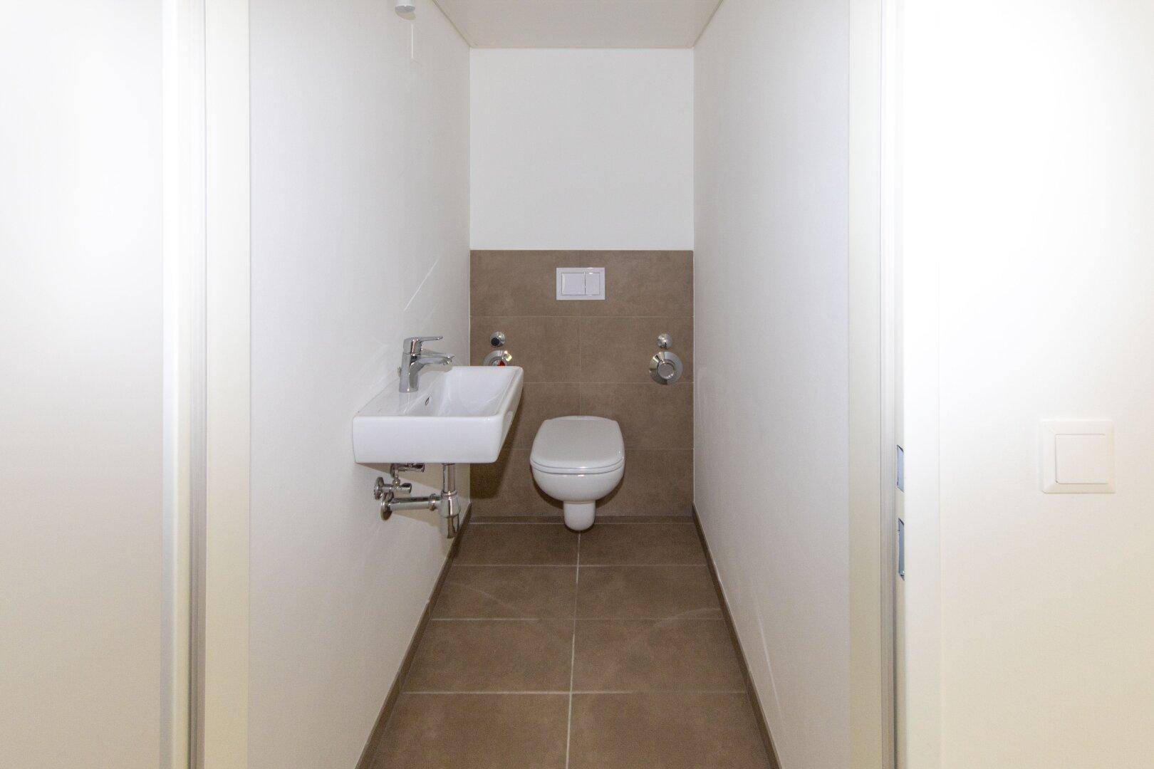Kompaktes Gäste-WC mit wandhängender Toilette, kleinem Waschbecken und modernen Fliesen.