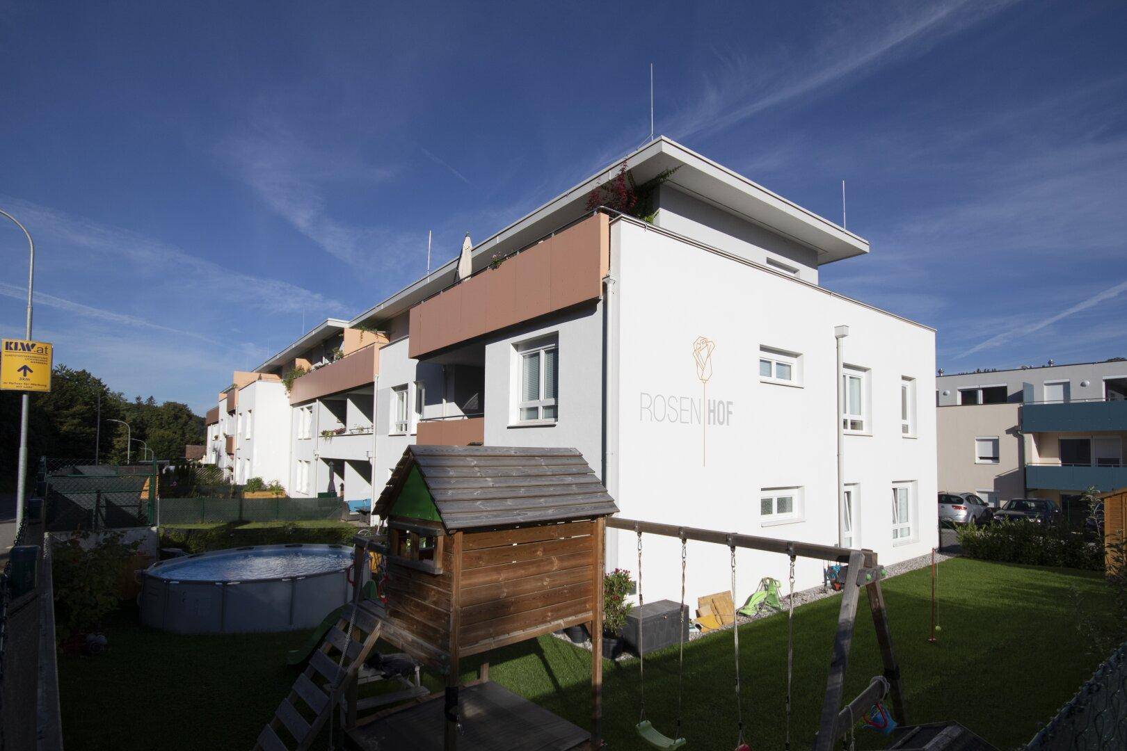 Modernes Mehrfamilienhaus mit Balkonen, umgeben von einem Spielplatz und einem Aufstellpool.