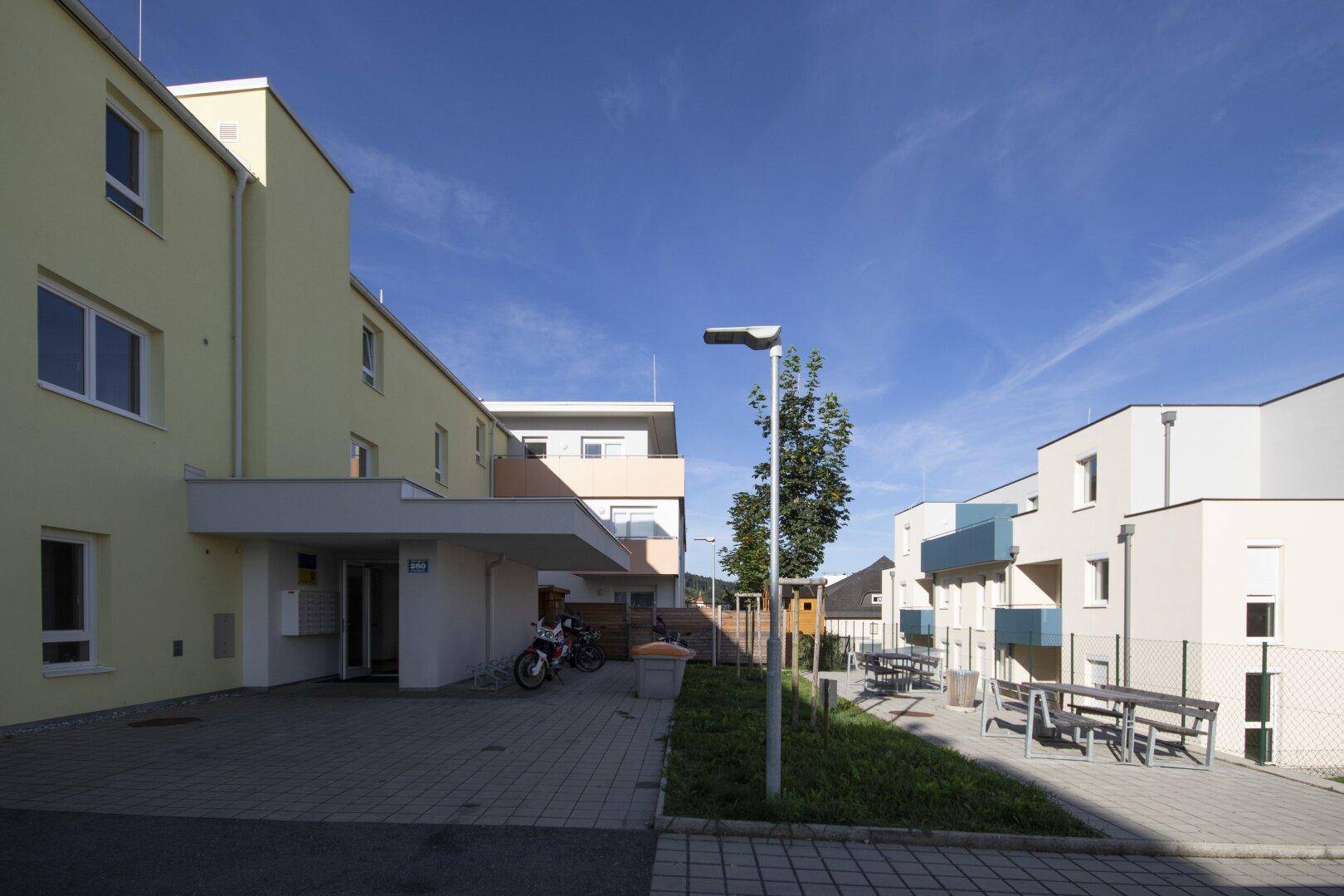 Eingangsbereich des Wohngebäudes mit überdachtem Zugang, Parkplätzen und Gemeinschaftsbereich im Freien.