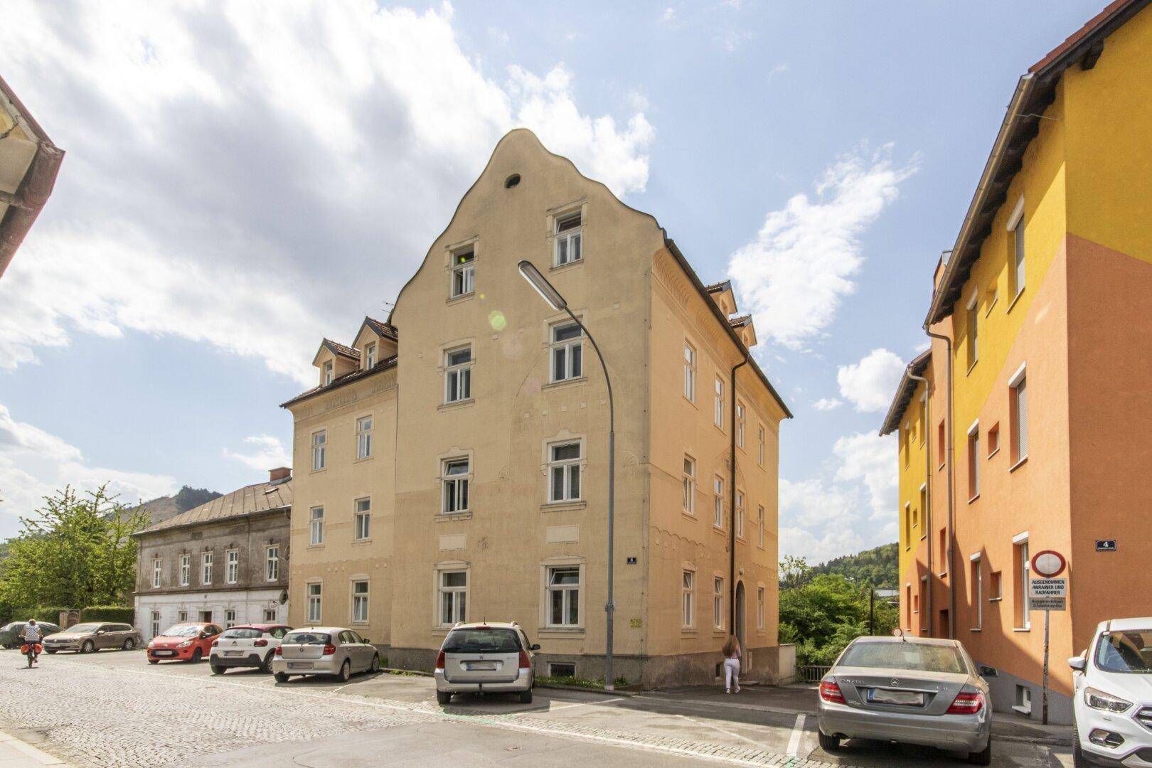 Historisches Wohngebäude mit markantem Giebel und Parkplätzen an der Straße, umgeben von grünen Hügeln.