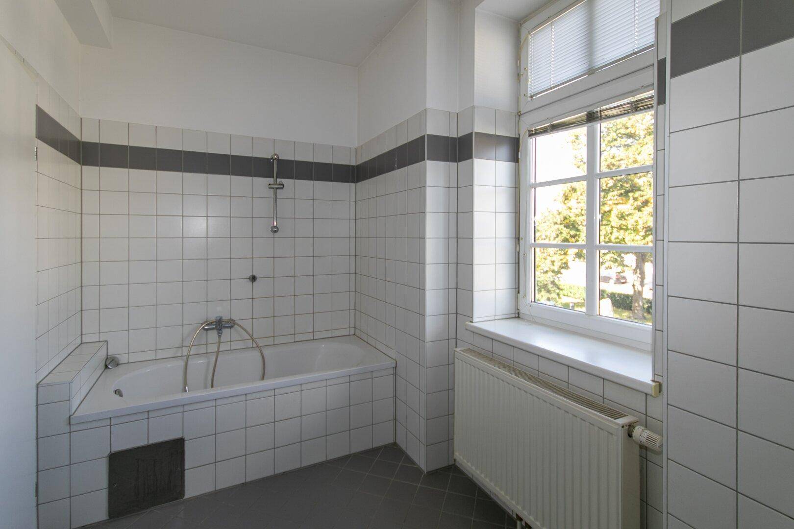 Gefliestes Badezimmer mit Badewanne, Fenster und Heizkörper, bereit zur Nutzung.