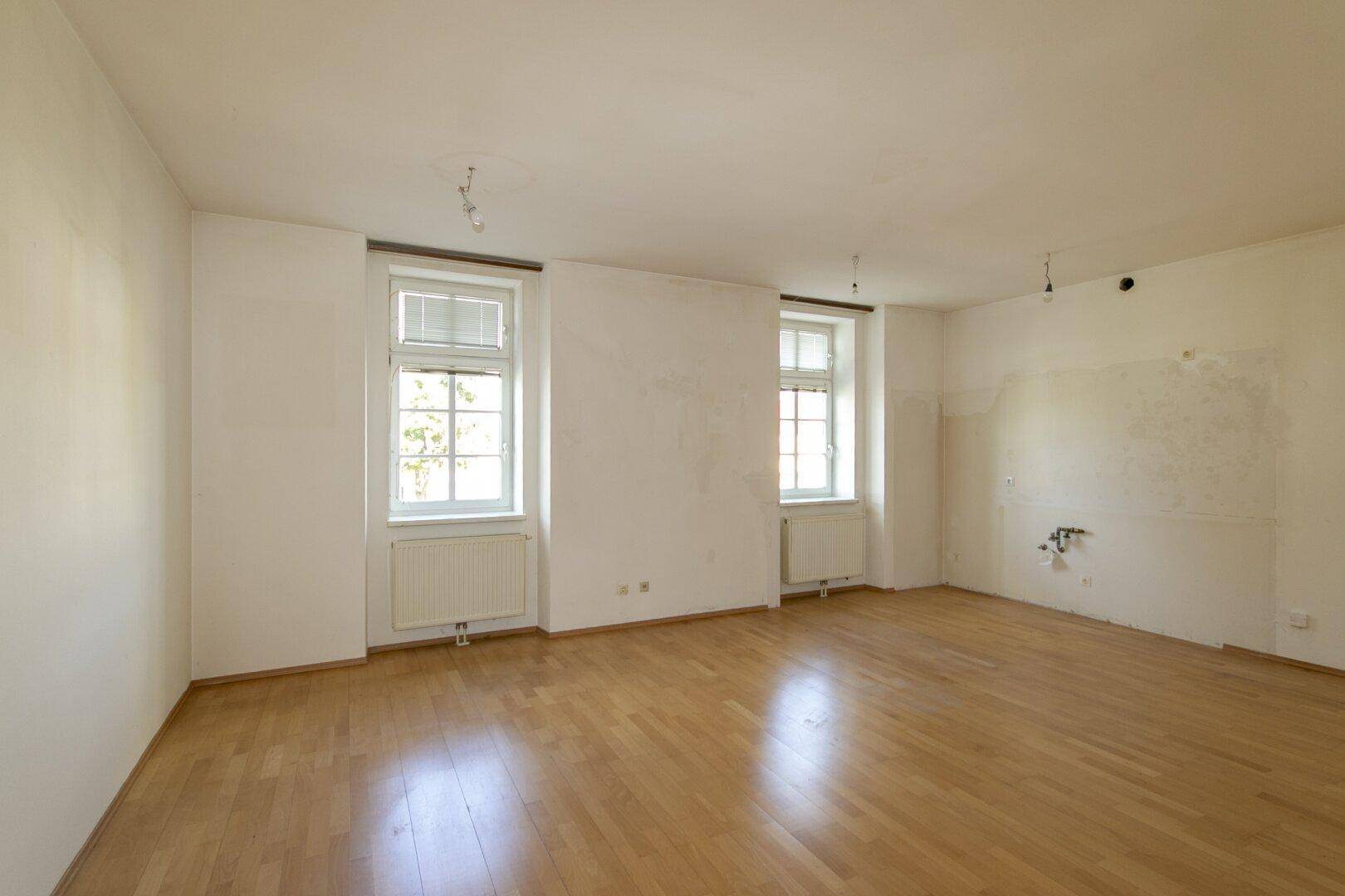 Heller Raum mit Holzfußboden, zwei Fenstern und Anschlüssen an der Wand.