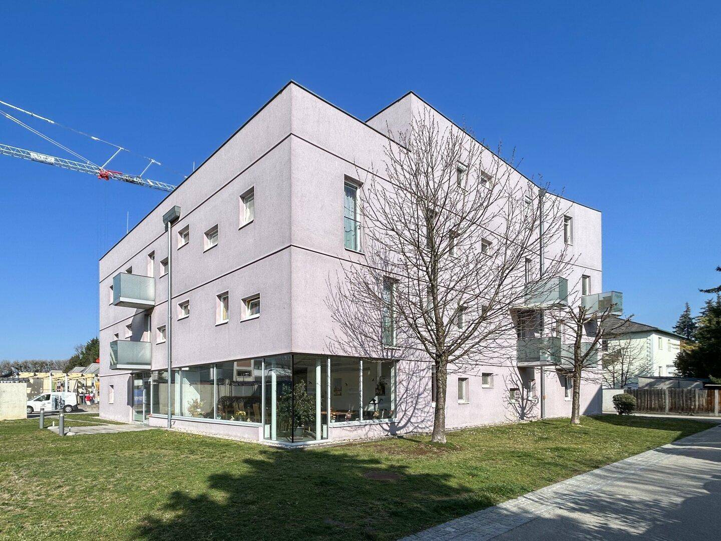 Modernes Gebäude mit heller Fassade, großen Fenstern im Erdgeschoss und kleinen Balkonen, umgeben von Grünflächen.