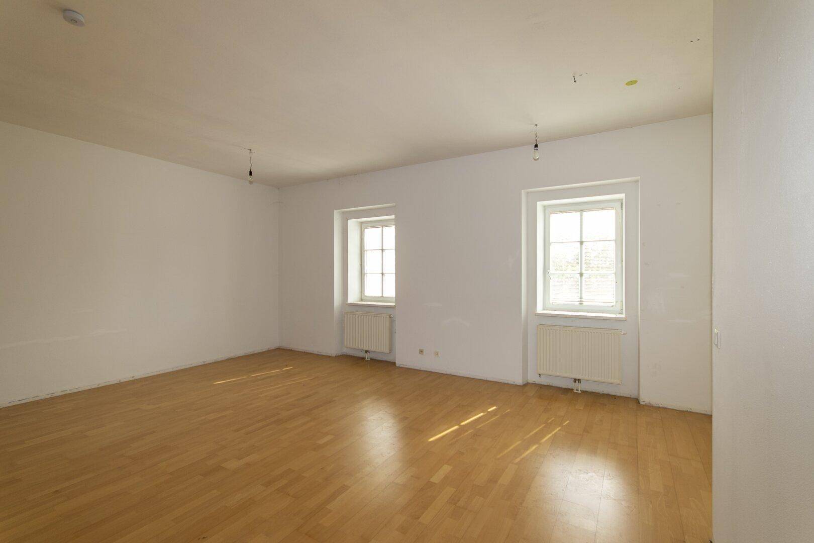 Großzügiger, heller Raum mit Parkettboden und zwei Fenstern, ideal für ein Wohnzimmer.