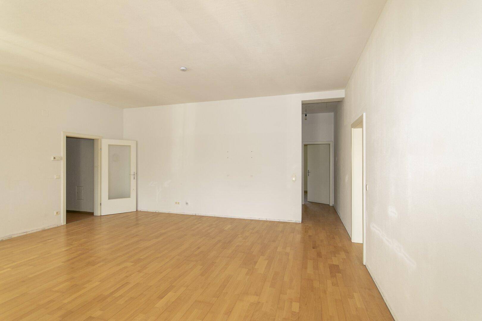 Geräumiges, leeres Zimmer mit hellem Parkettboden und Zugang zu weiteren Räumen.