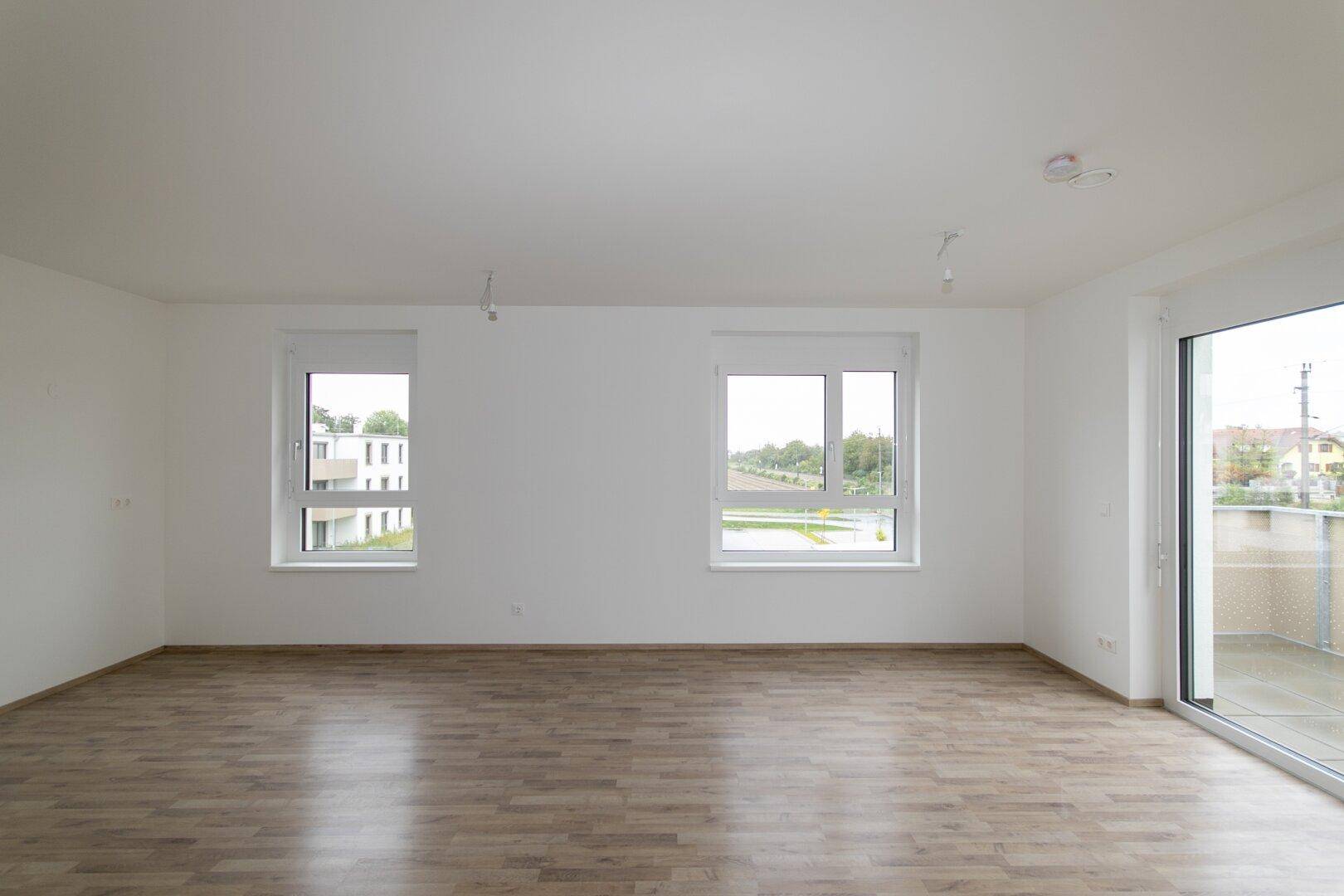 Geräumiges, helles Wohnzimmer mit Laminatboden und großen Fenstern, bietet viel Tageslicht.
