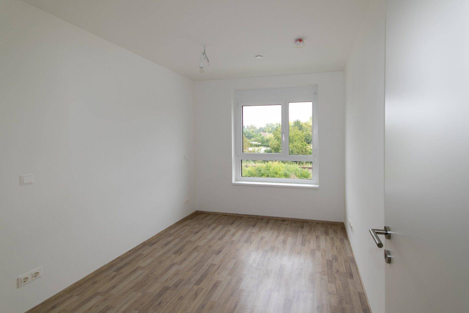 Helles Schlafzimmer mit Laminatboden und Fenster mit Blick ins Grüne, unmöbliert.