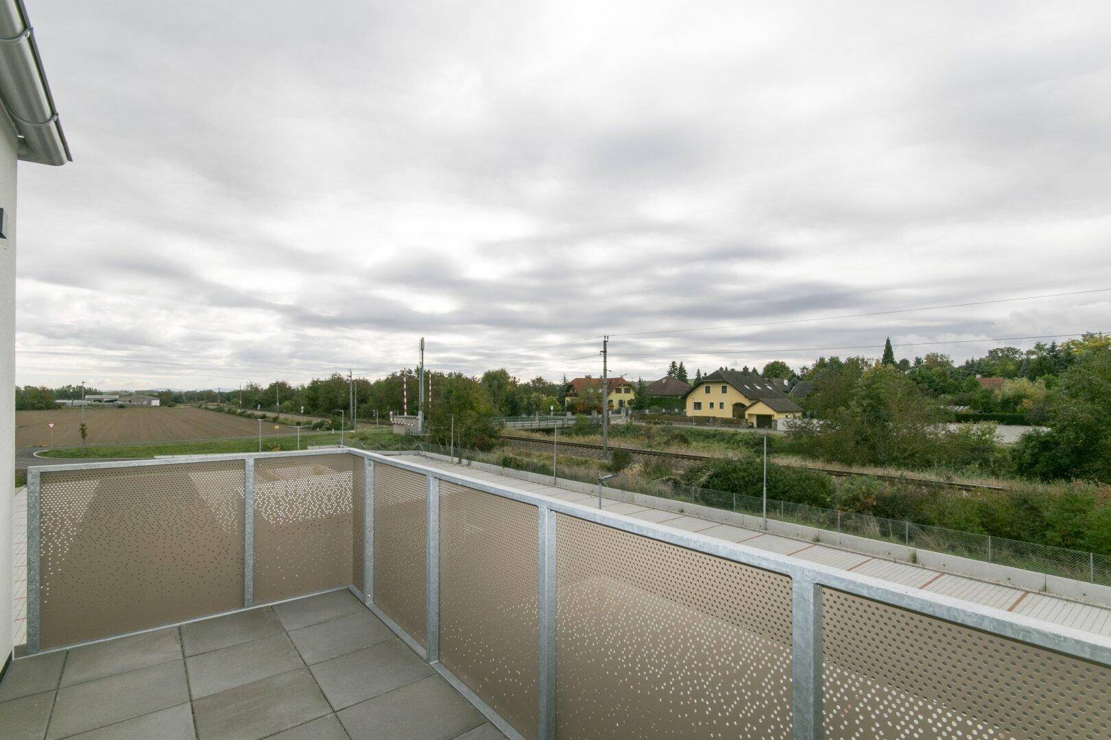 Balkon mit Fliesenboden und moderner Lochblechbrüstung, Blick auf Felder und Bahngleise.