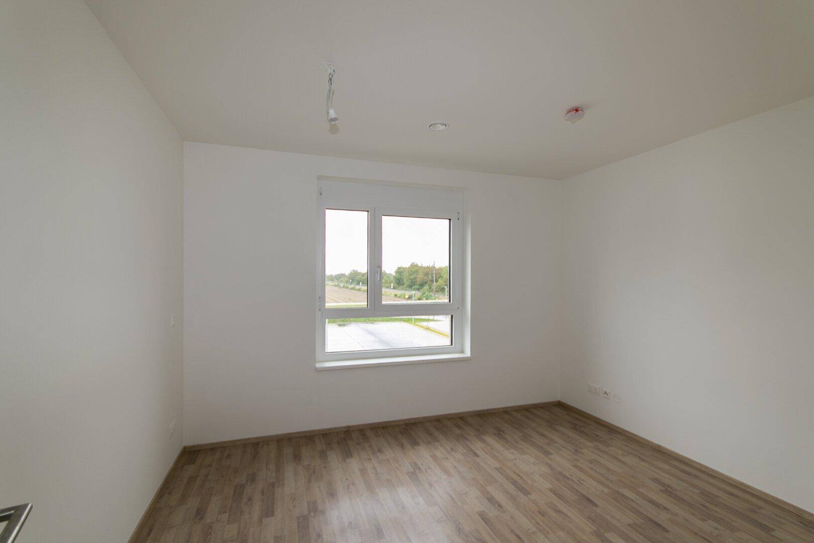 Unmöbliertes Schlafzimmer mit Laminatboden und großem Fenster, das einen Blick ins Grüne bietet.