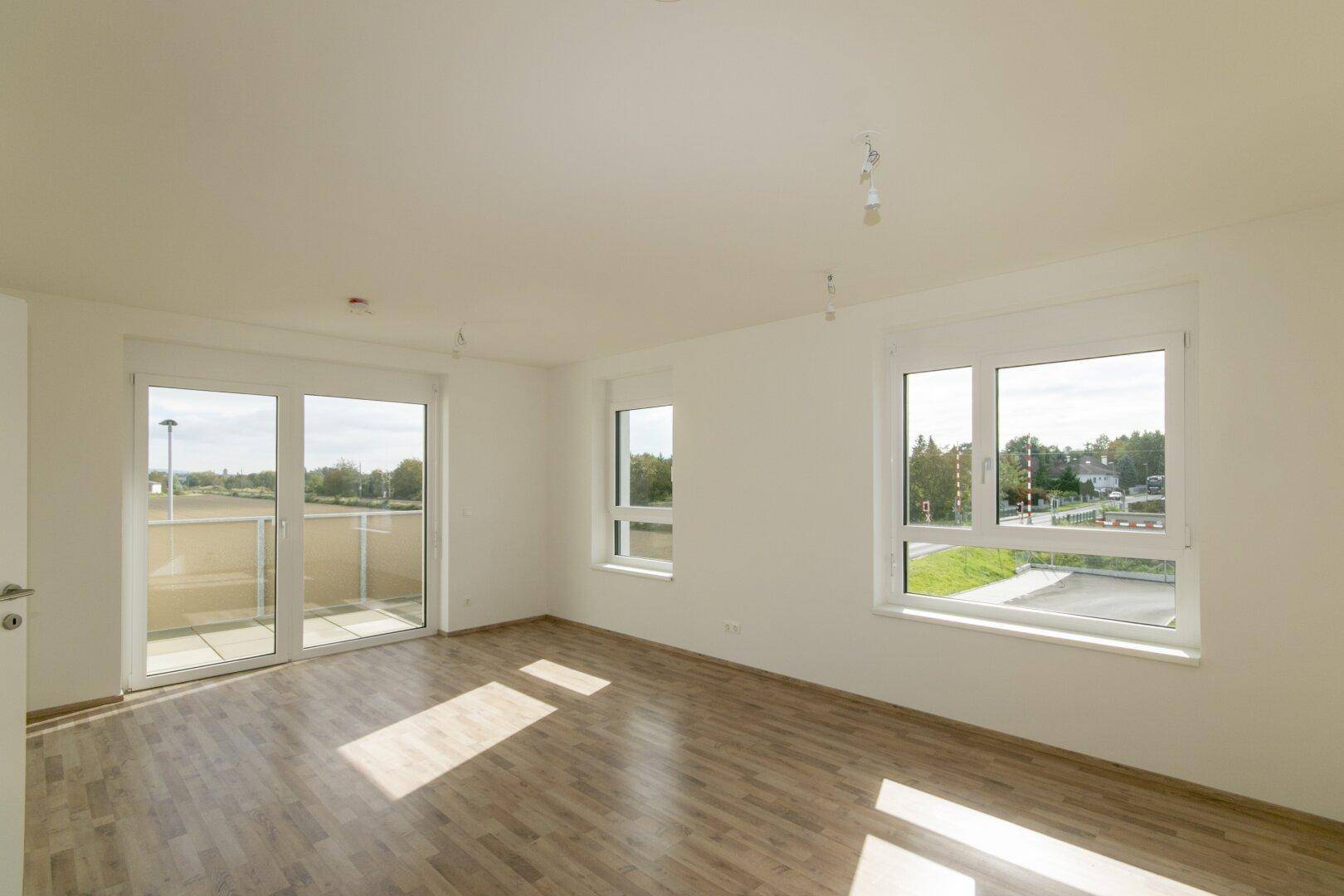 Geräumiger, heller Raum mit Laminatboden, großen Fenstern und Zugang zum Balkon.