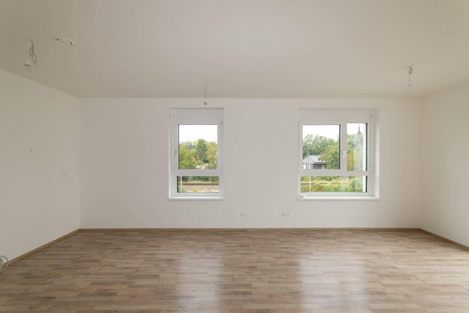 Geräumiges, unmöbliertes Zimmer mit Laminatboden und zwei großen Fenstern mit Blick ins Grüne.