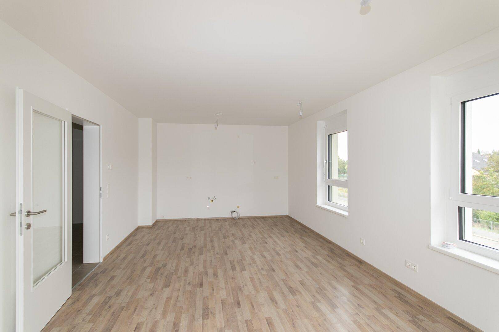Leerer Raum mit Laminatboden und Anschlüssen für eine Küche, hell durch große Fenster.