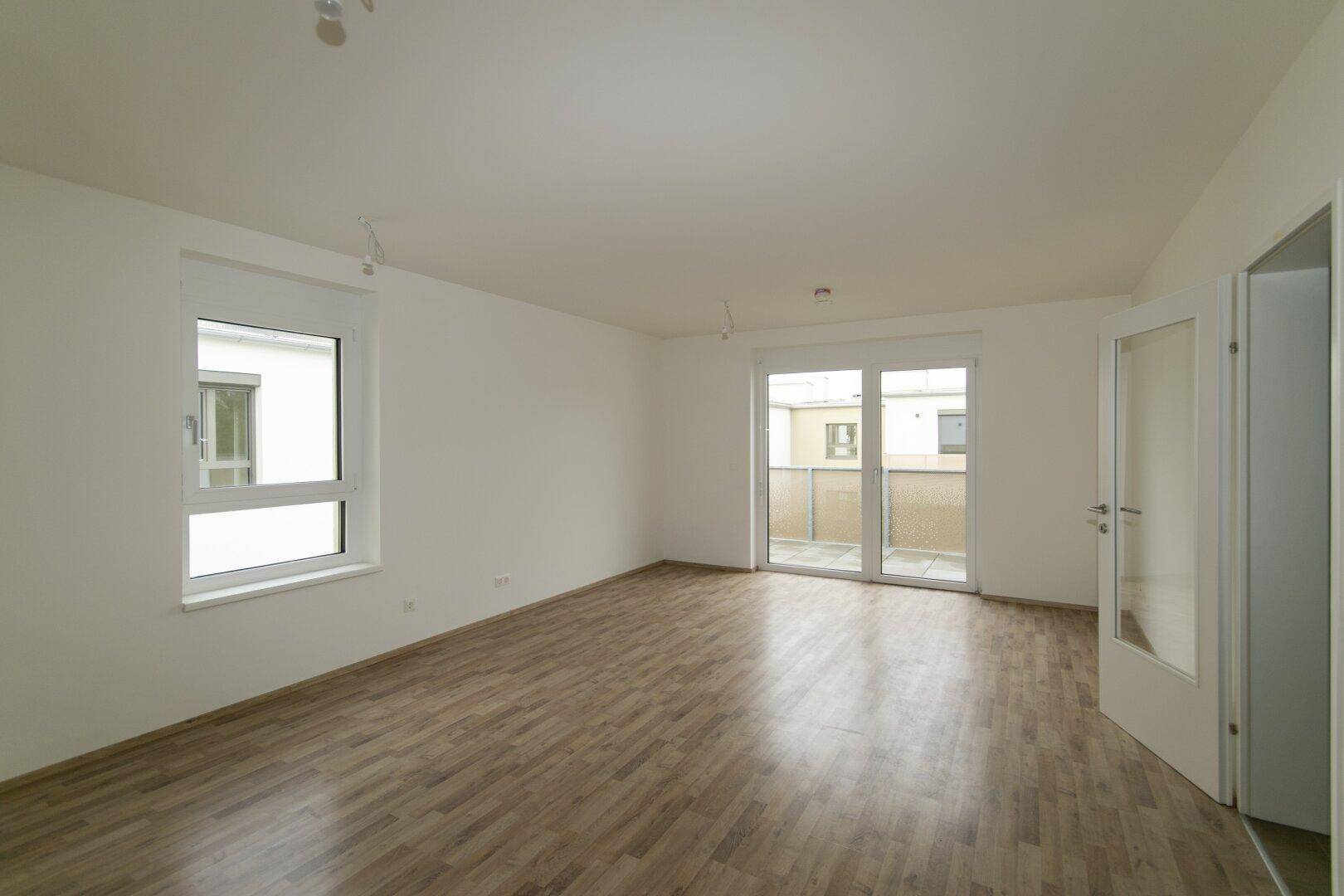 Leeres Wohnzimmer mit Laminatboden, großem Fenster und Zugang zu einem Balkon.