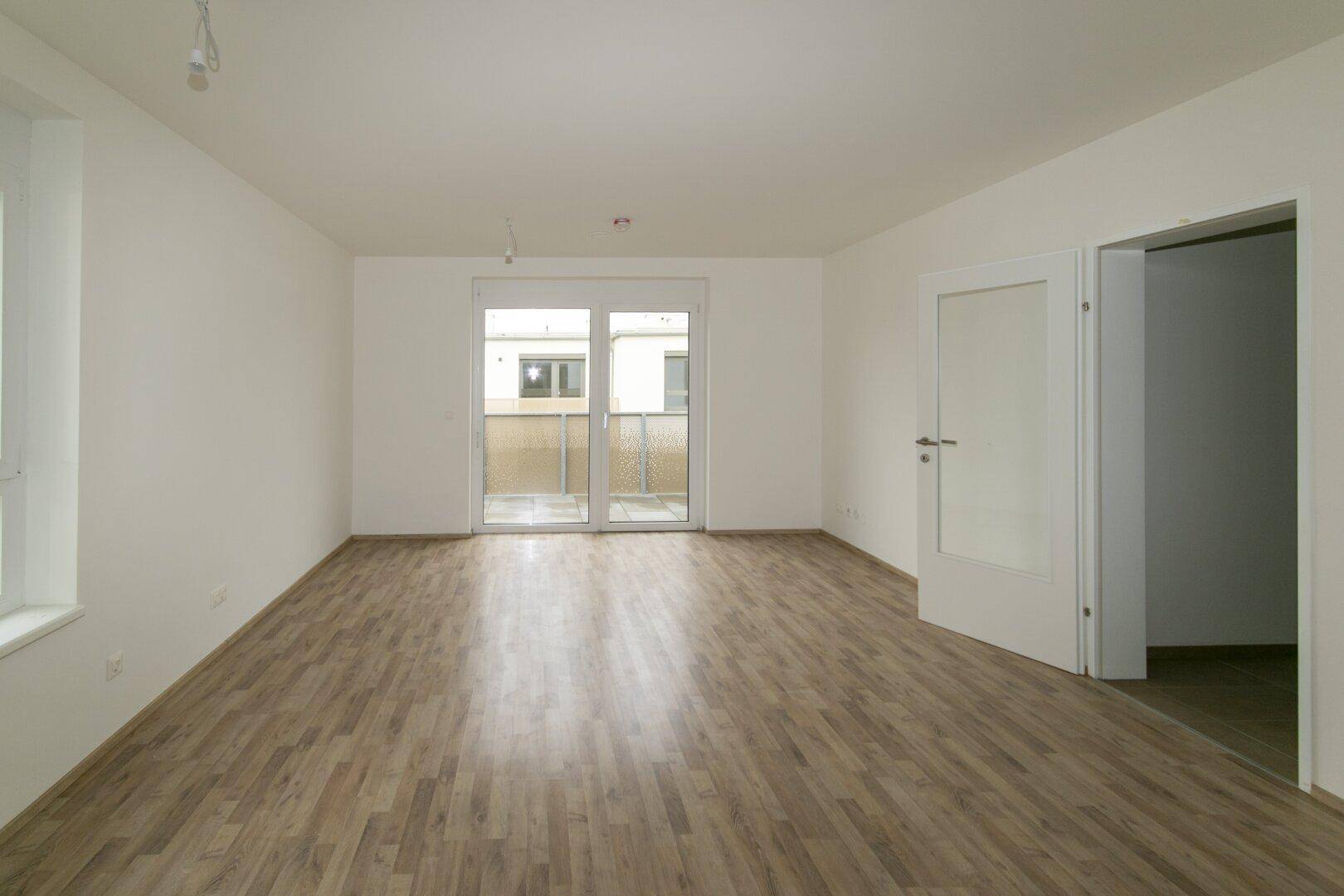 Helles Wohnzimmer mit Laminatboden und breiter Schiebetür, die auf den Balkon führt.