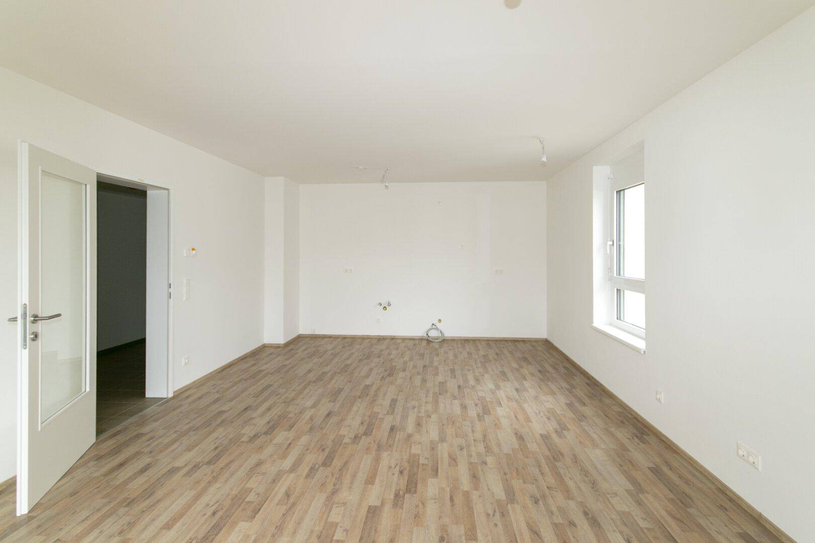 Geräumiges, leeres Wohnzimmer mit Laminatboden und einem großen Fenster, das viel Licht spendet.
