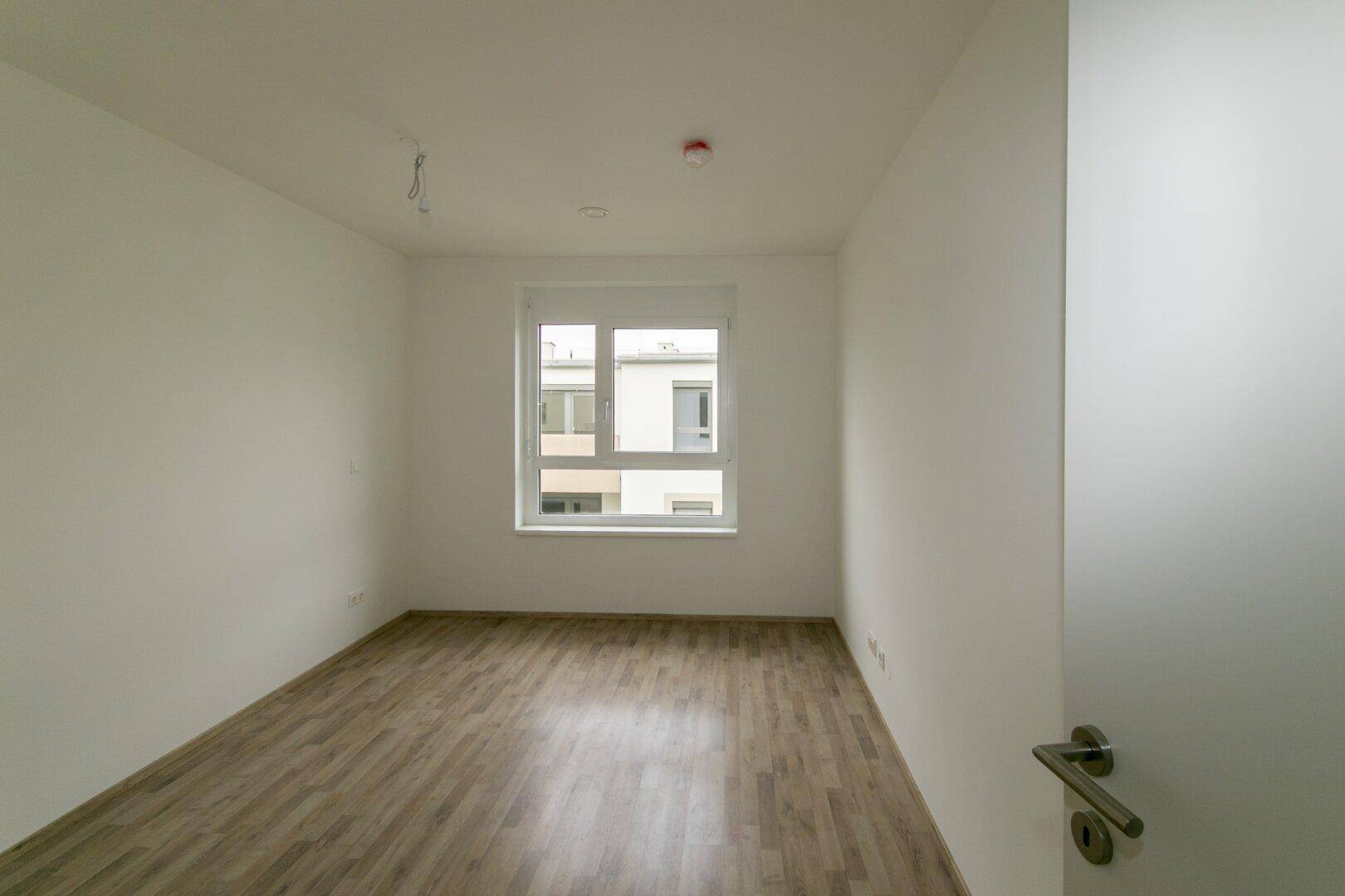 Leeres Schlafzimmer mit Laminatboden und einem Fenster mit Blick auf die Nachbargebäude.