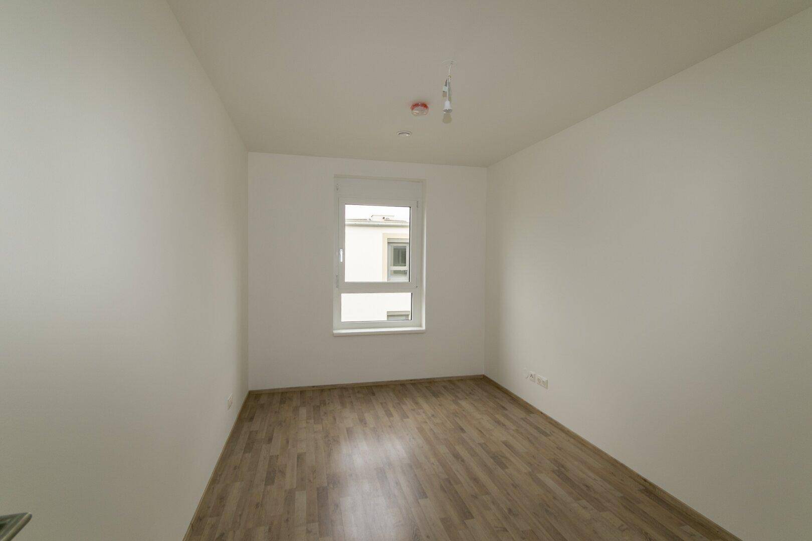 Helles Schlafzimmer mit Laminatboden und einem Fenster, das viel Tageslicht hereinlässt.