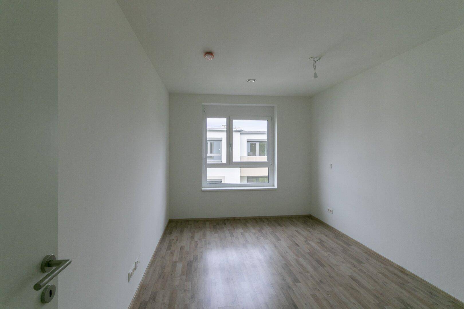 Helles Schlafzimmer mit Laminatboden und Fenster, das einen Blick auf die Nachbargebäude bietet.