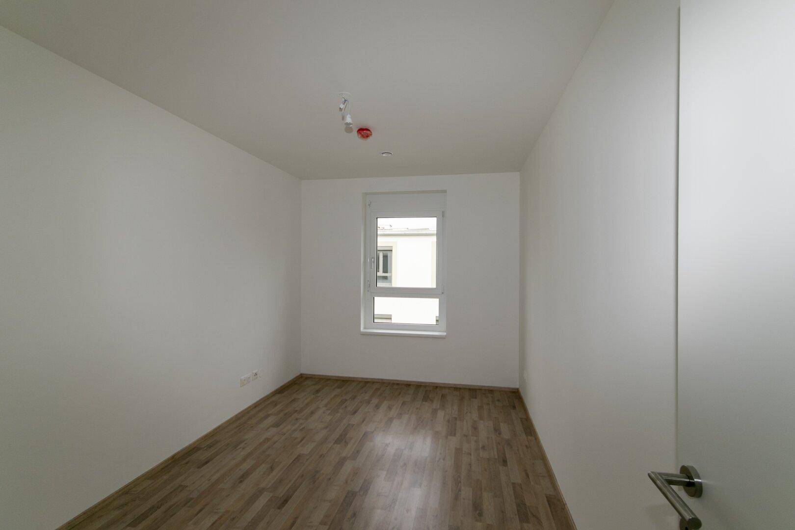 Leeres Schlafzimmer mit Laminatboden und einem Fenster, das viel Tageslicht hereinlässt.
