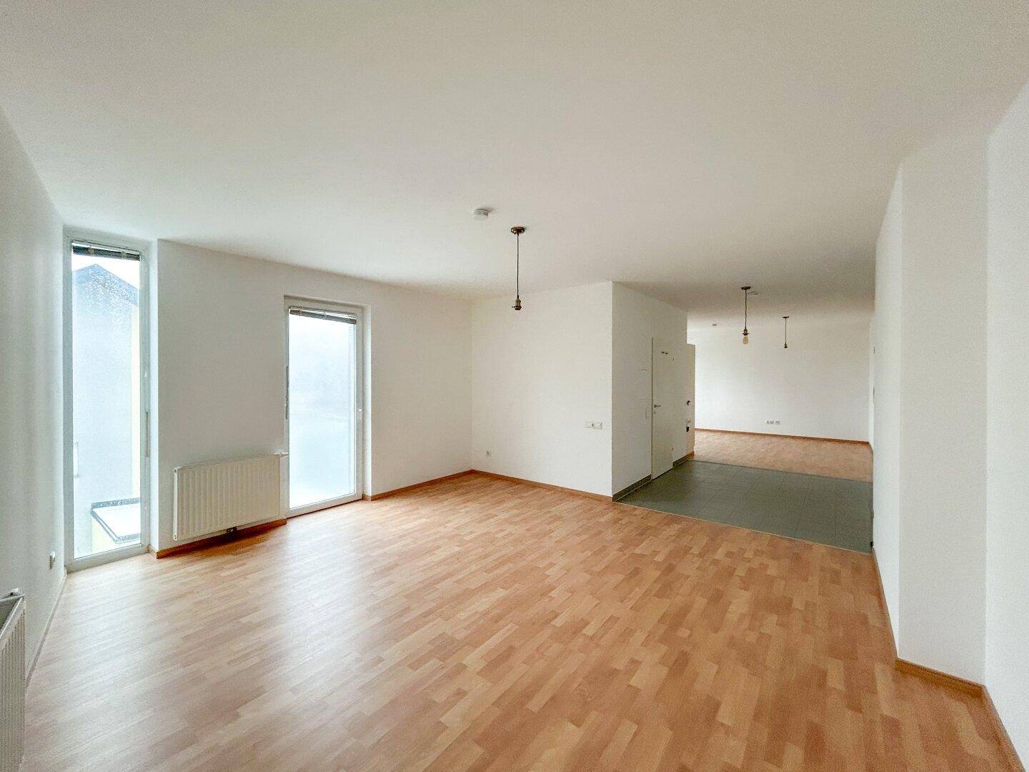 Offener Wohnbereich mit Laminatboden, weißen Wänden und großem Fenster.