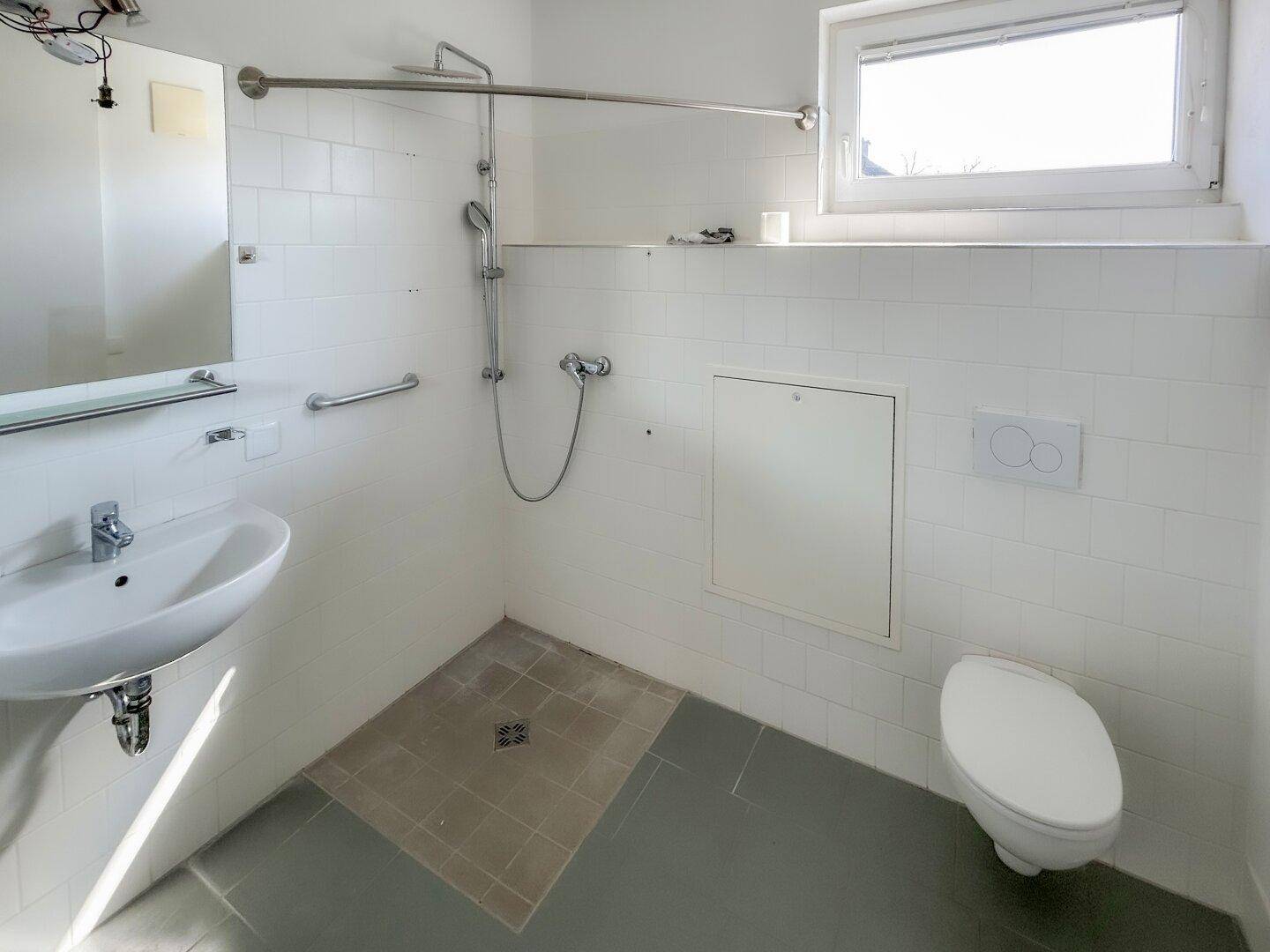 Funktionales Badezimmer mit Dusche, WC, Waschbecken, Fenster und Haltegriffen.