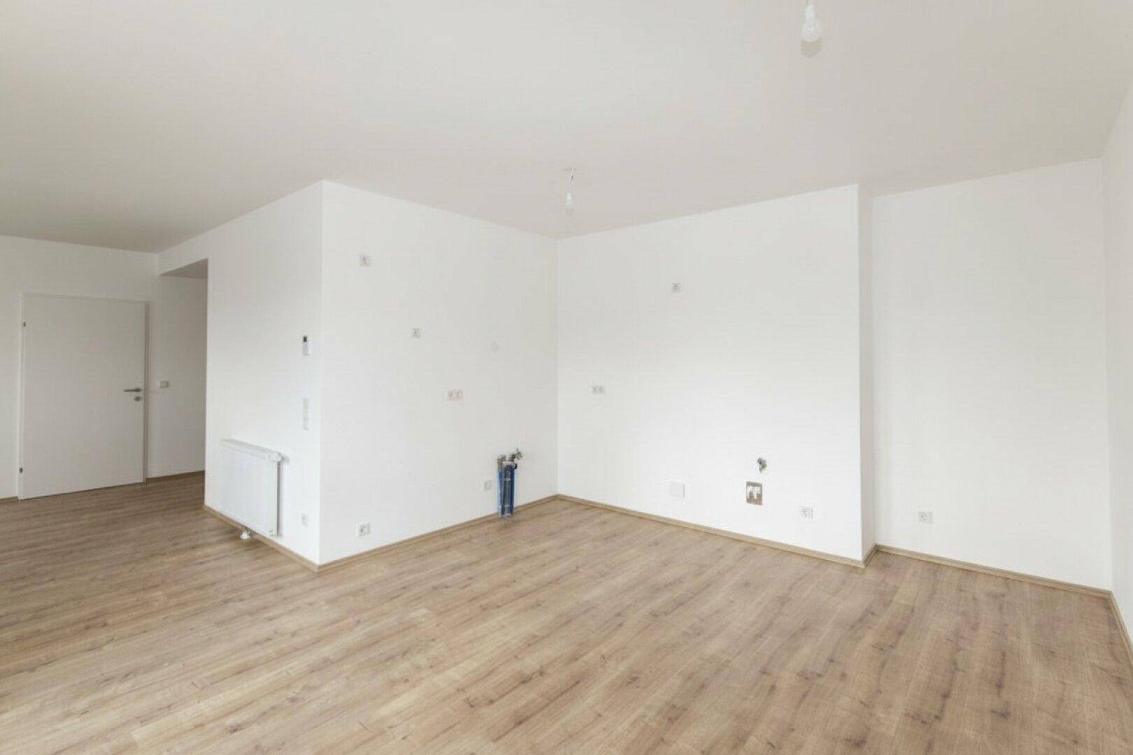 Leerer Raum mit Laminatboden und weißen Wänden, vorbereitet für die Installation einer Küche.